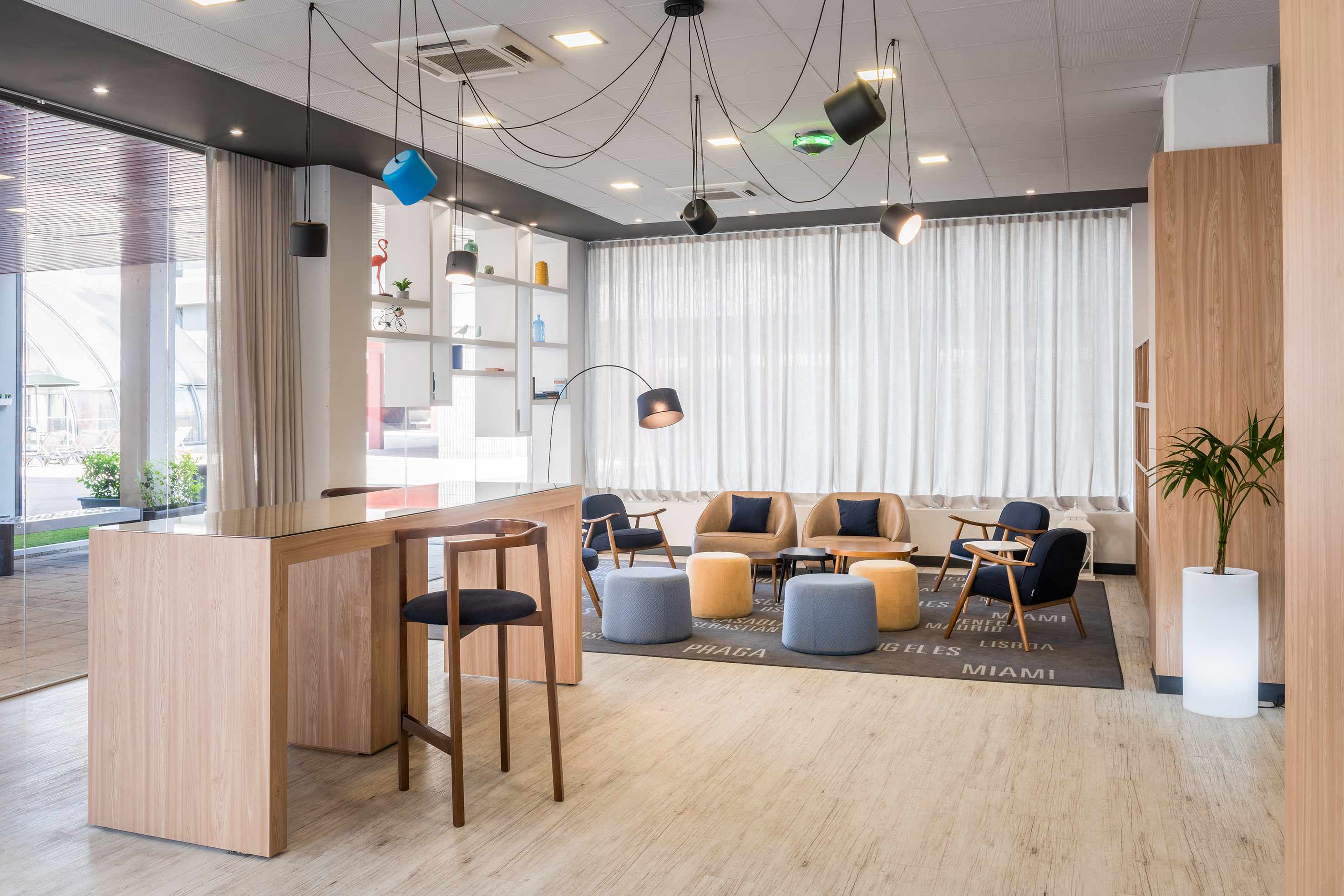Modern hotel lounge with wooden bar, armchairs, colorful ottomans, pendant lights.