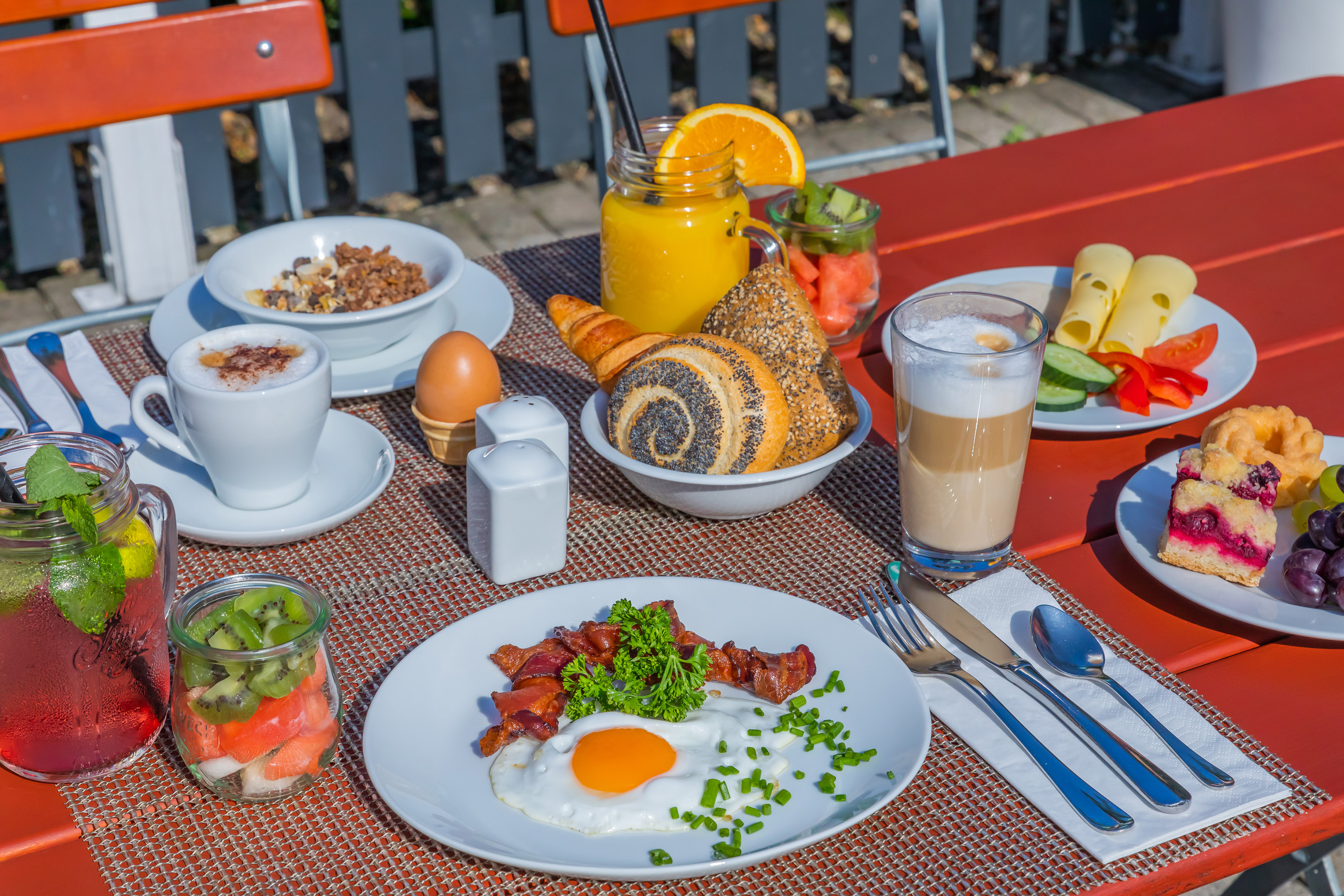 Outdoor breakfast spread: fried egg, bacon, coffee, juice, pastries, fruit salad.