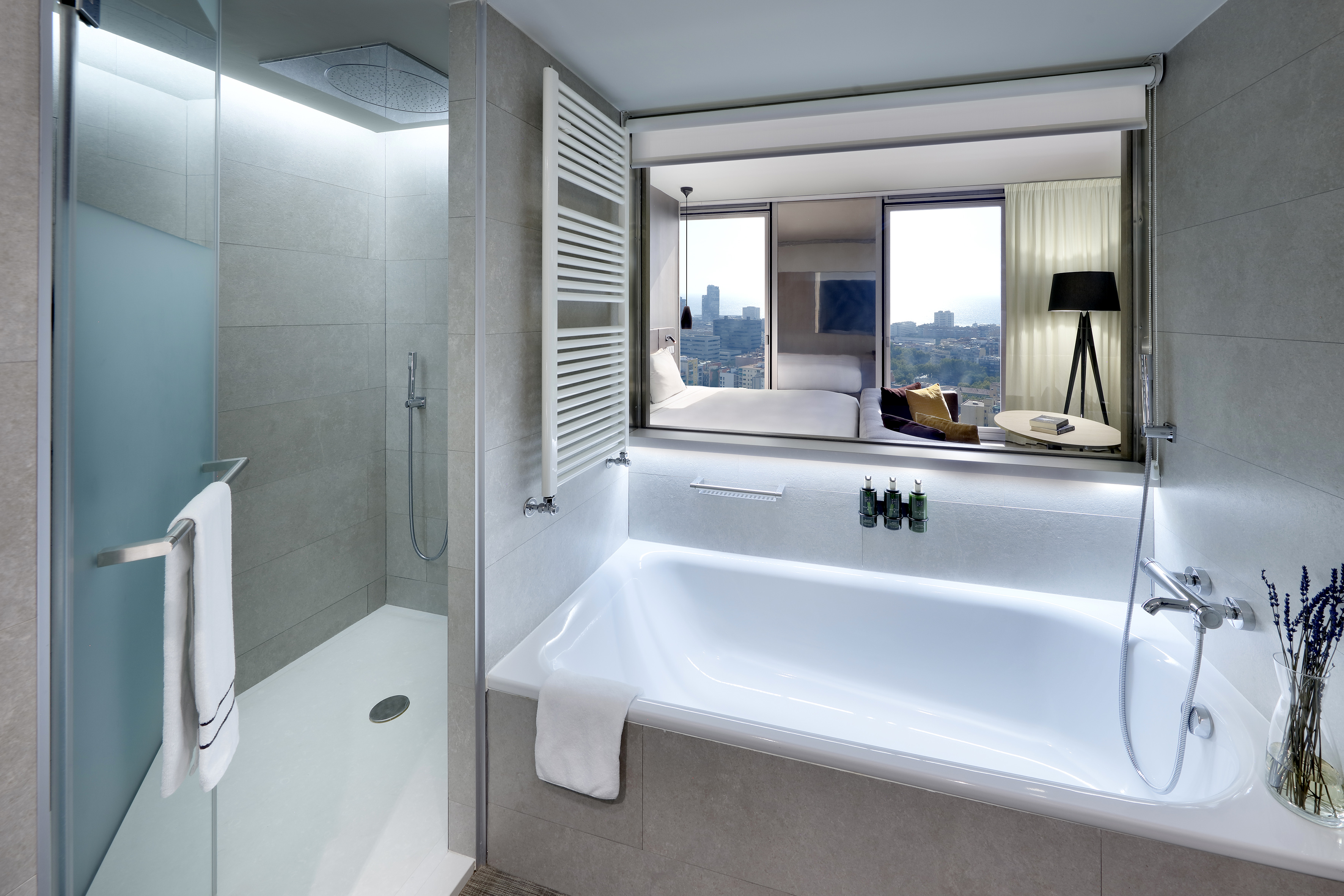 Modern hotel suite bathroom with bathtub, shower, and bedroom city view.