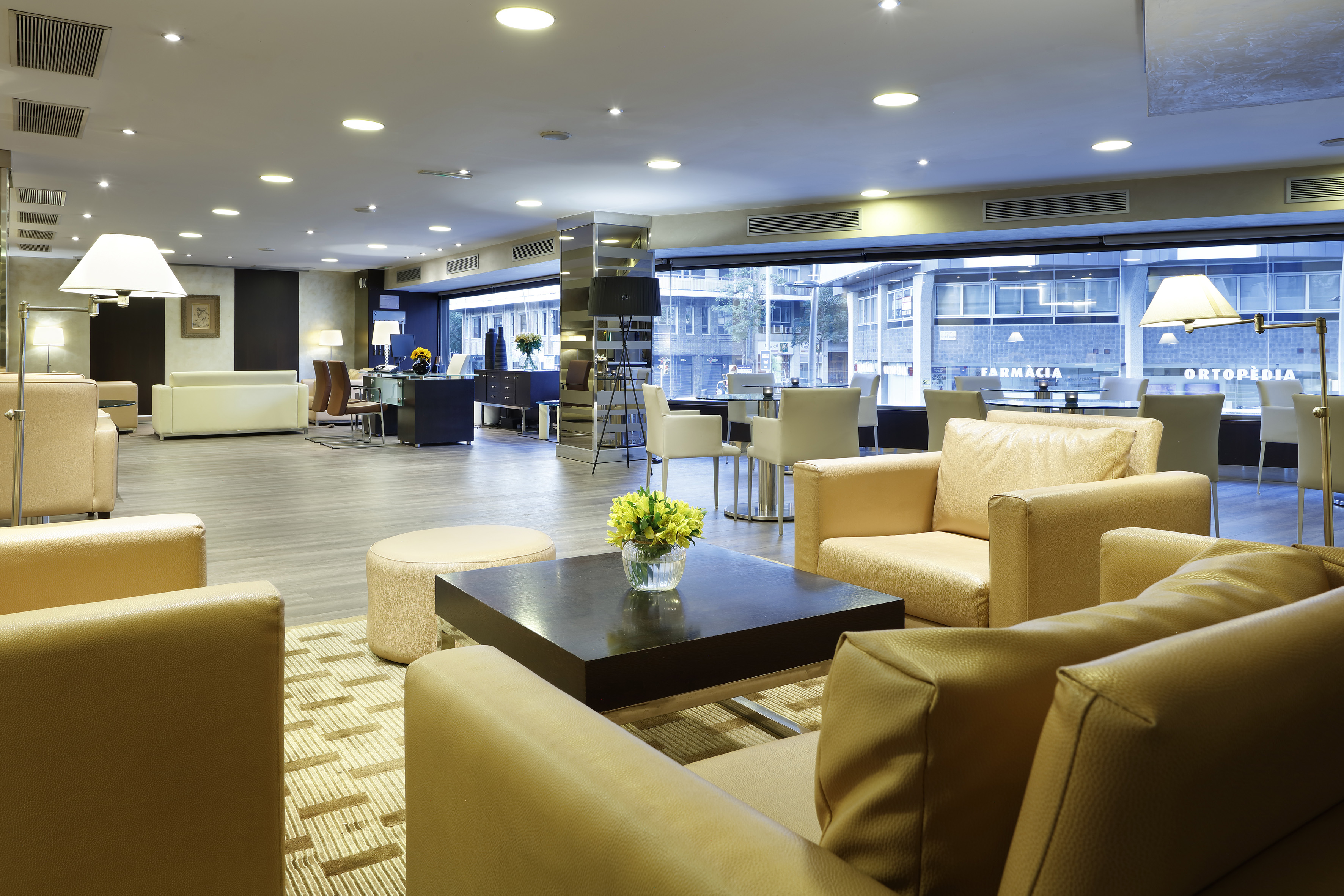 Modern hotel lounge with beige leather seating, dark tables, and street view.