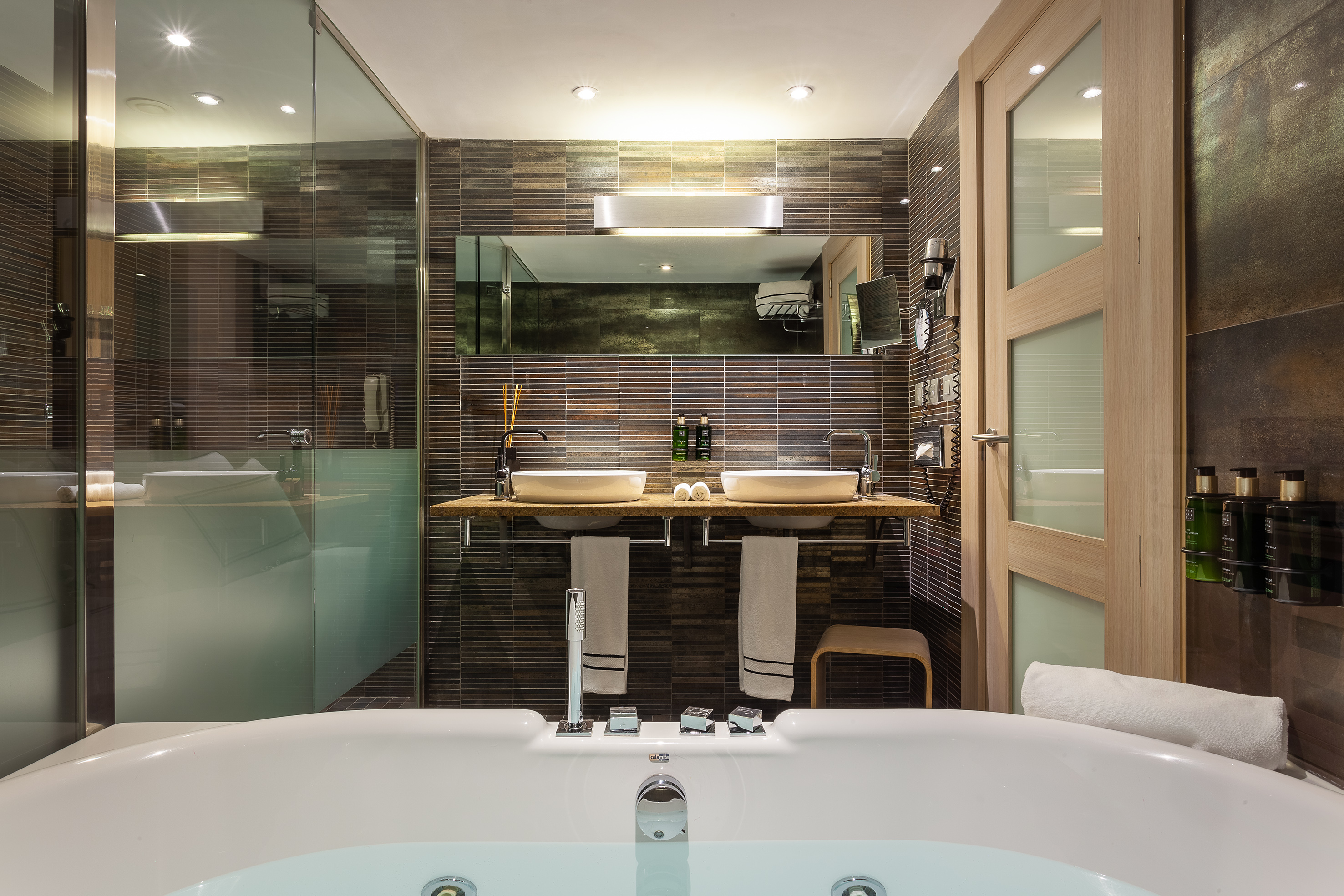 Modern hotel bathroom, large bathtub, dual vessel sinks, dark tiled walls.