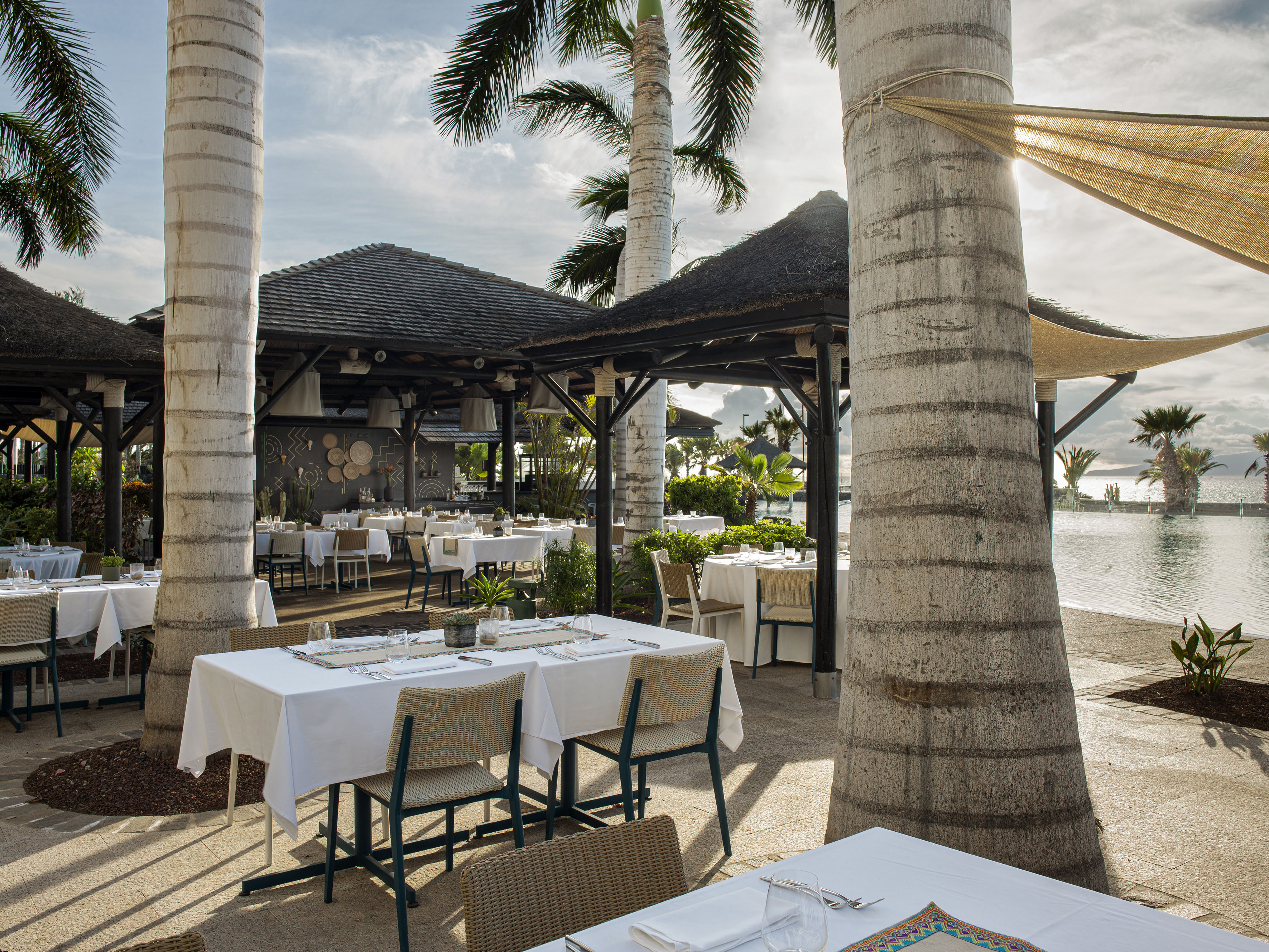 Tropical resort outdoor restaurant with dining tables, palm trees, and pool.