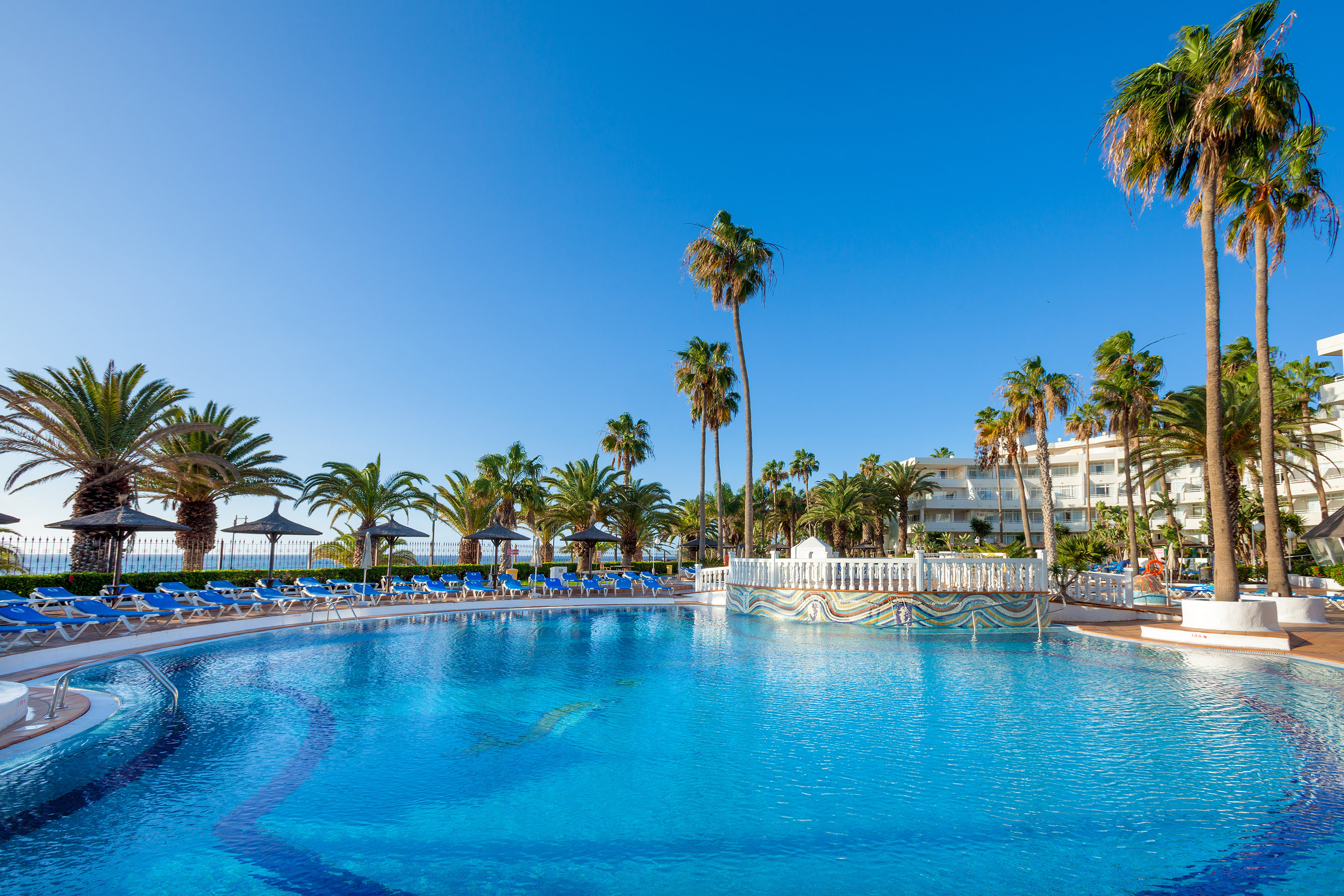 Sunny resort swimming pool with palm trees and blue loungers.