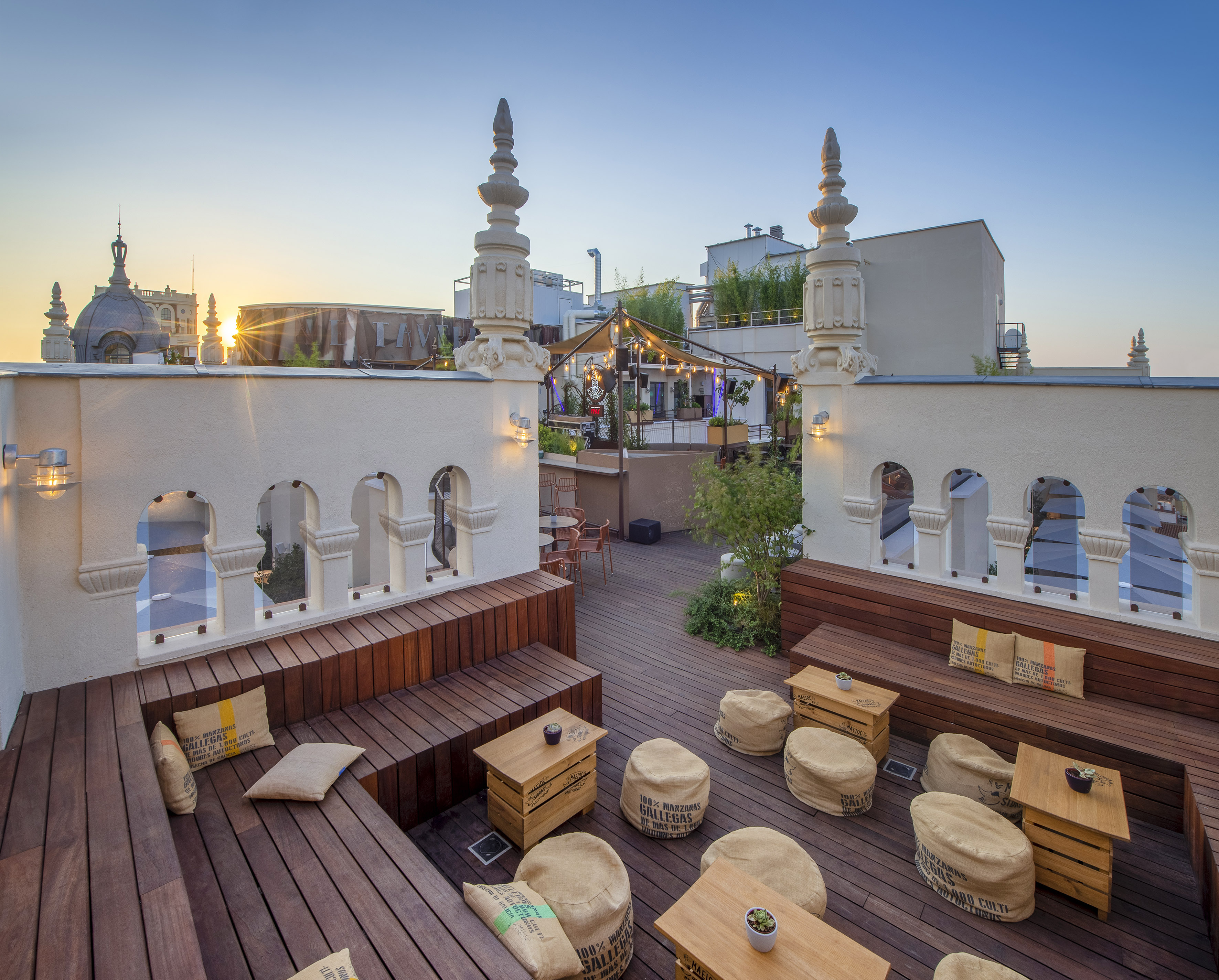 Rooftop bar at sunset with wooden seating, white arched wall, city view.