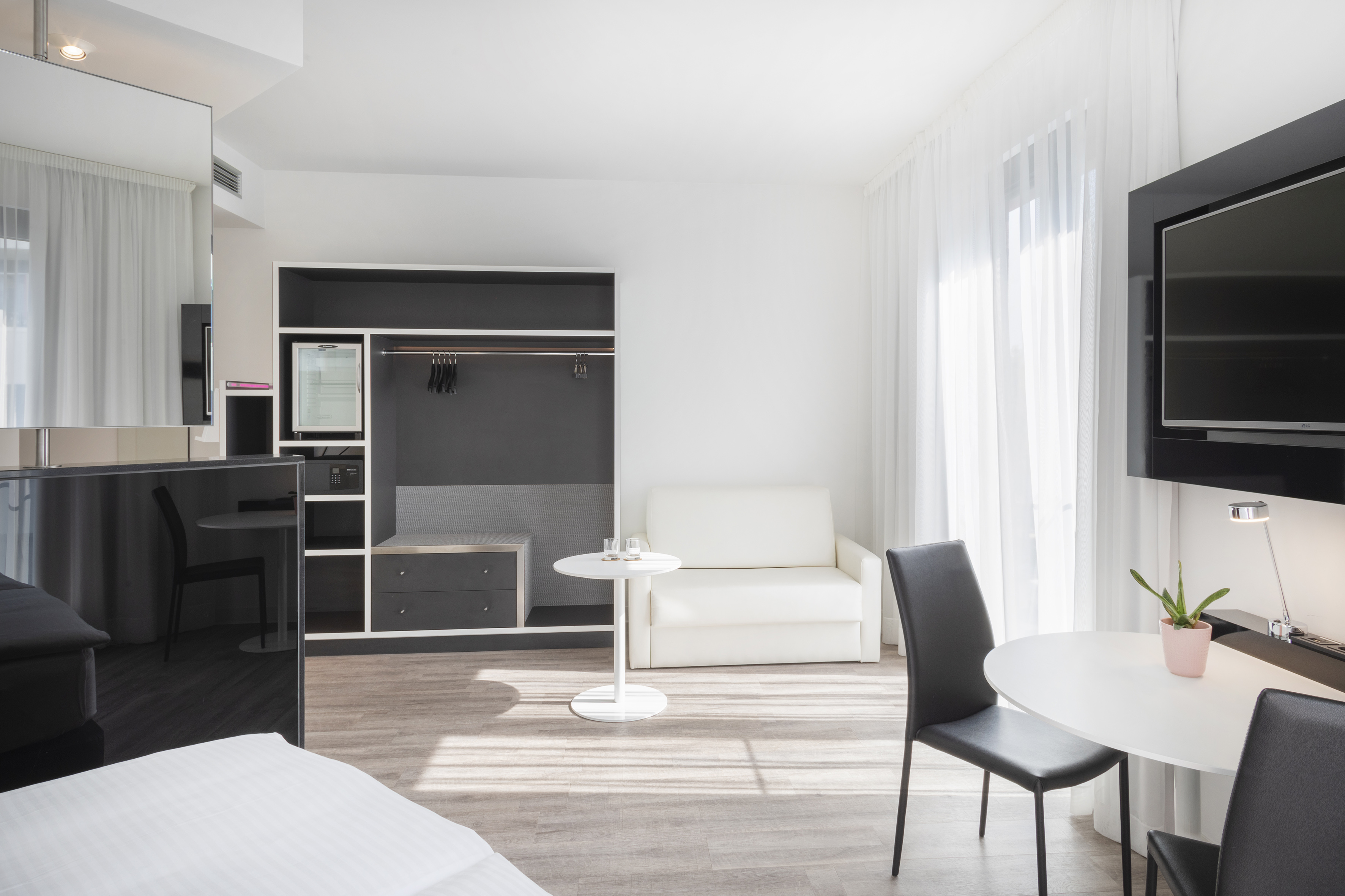 Bright, modern hotel suite with sofa, dining table, TV, and open wardrobe.