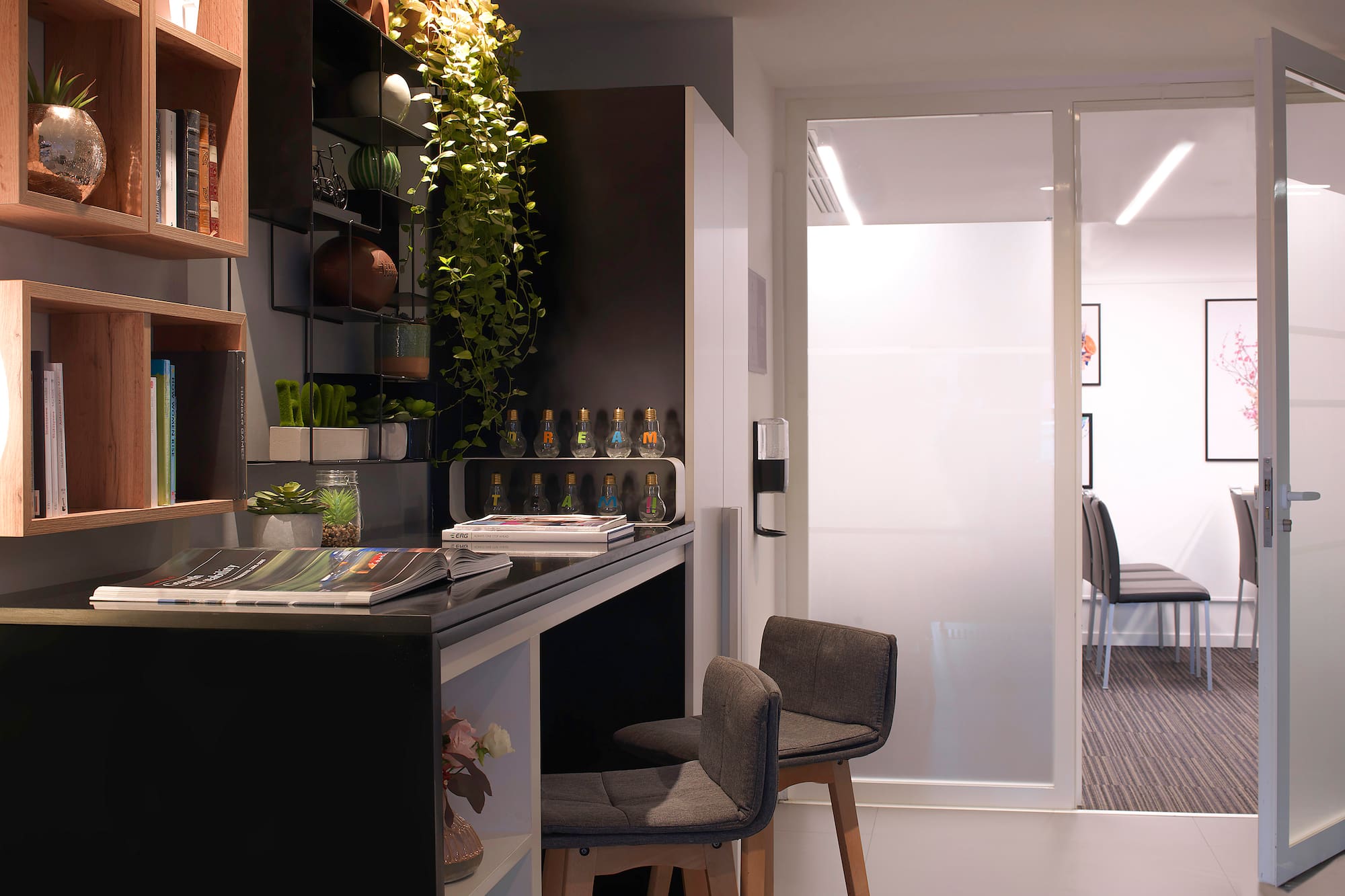 Modern office desk with bar stools, shelves, plants, and glass partition.