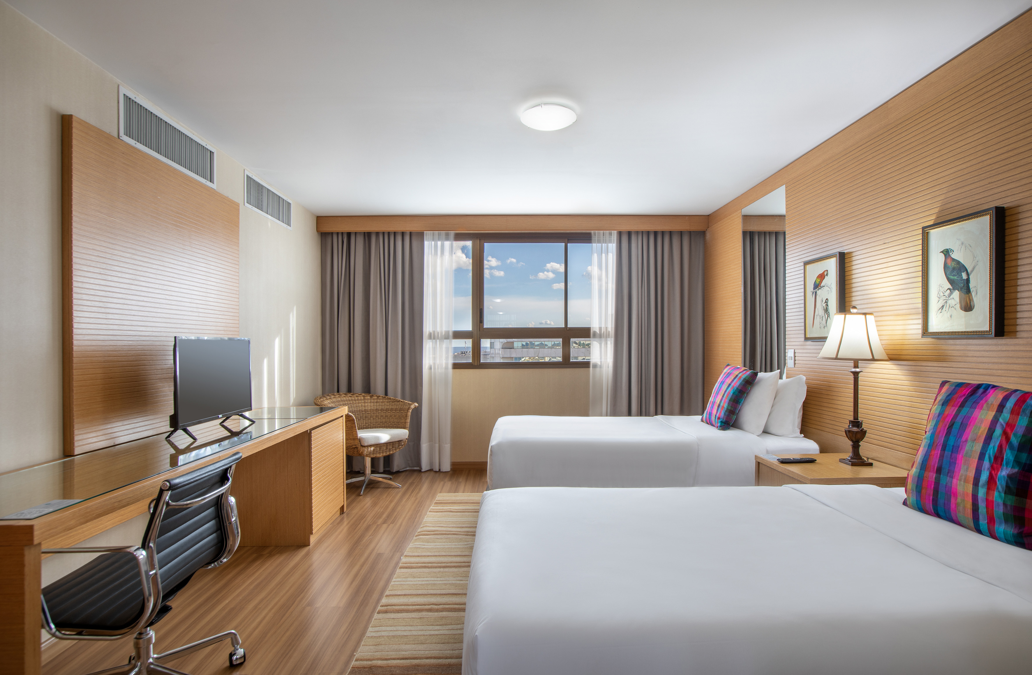 Modern hotel room with twin beds, desk, TV, and city view from window.