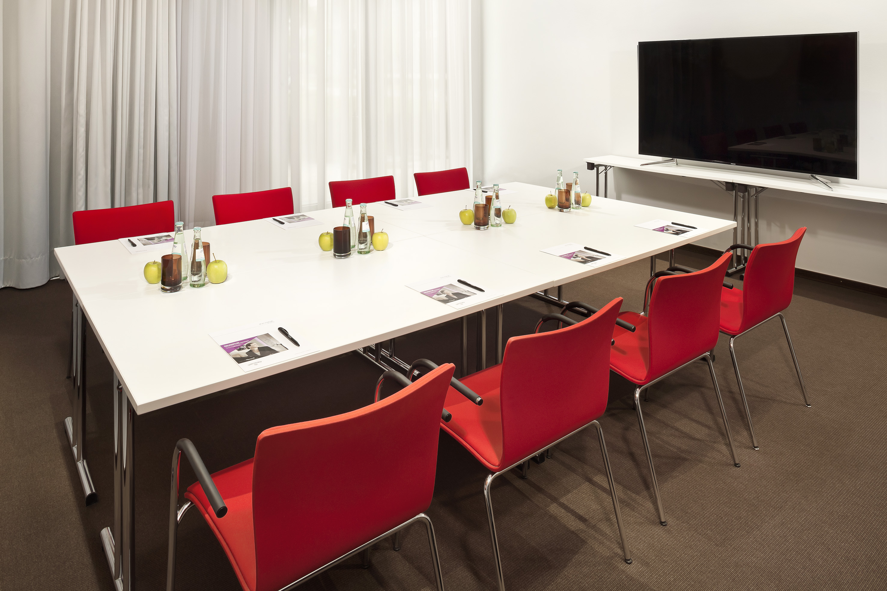 Modern conference room with long white table, red chairs, and TV monitor.