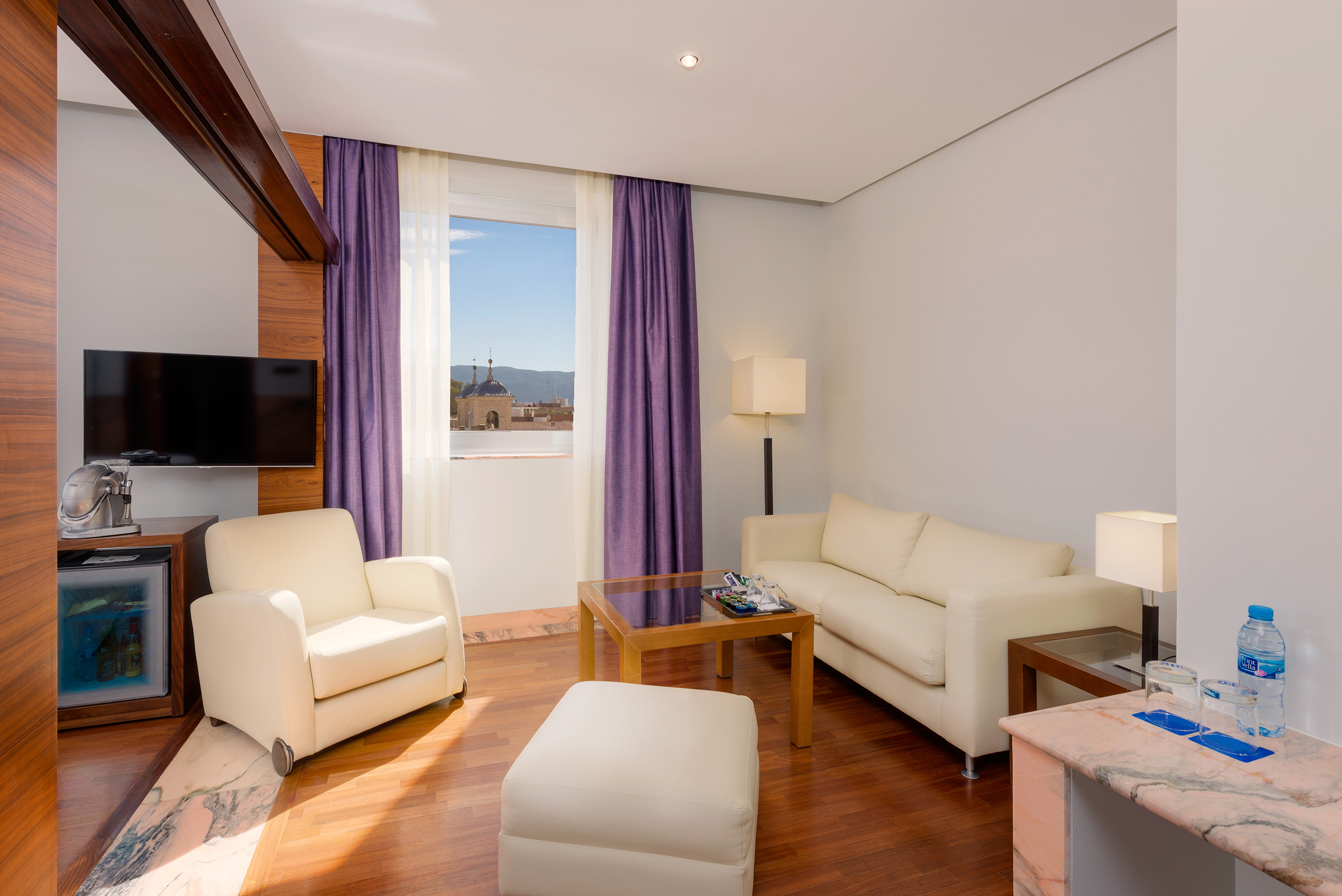 Modern hotel suite with cream seating, wooden floor, purple drapes, and city view.