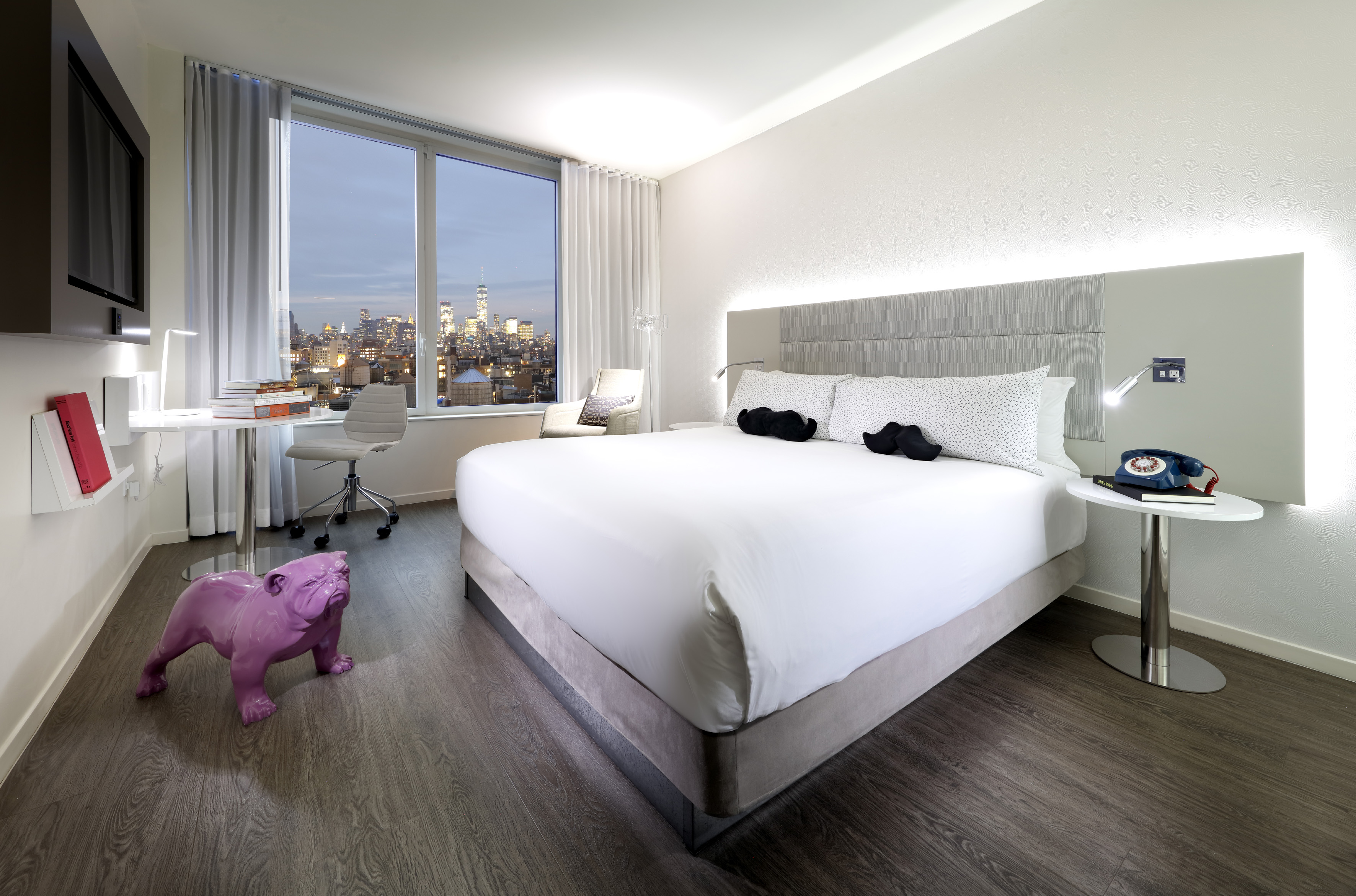 Hotel room with king bed, desk, NYC skyline view, and purple bulldog.