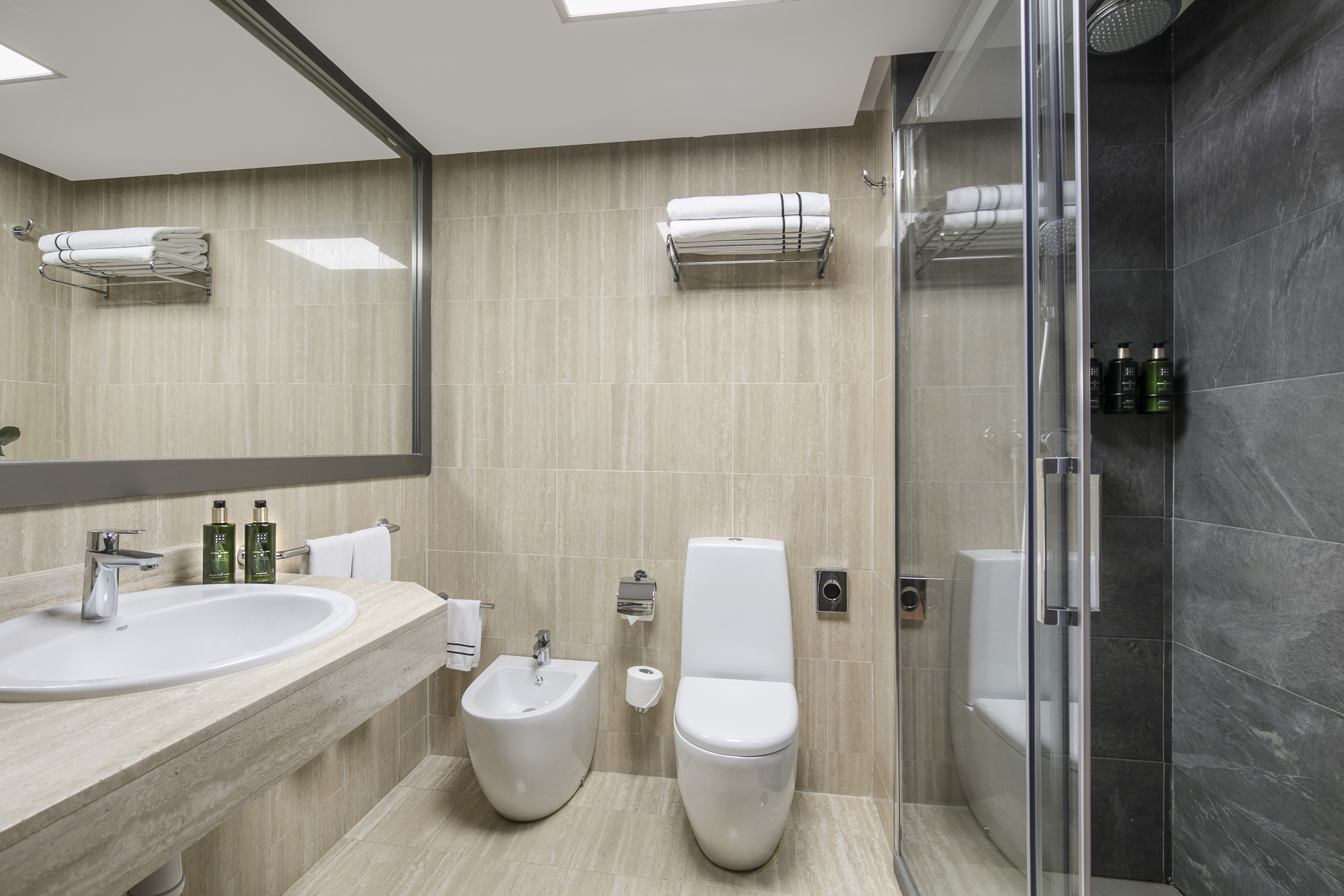 Modern hotel bathroom with beige tiles, sink, toilet, bidet, and glass shower.