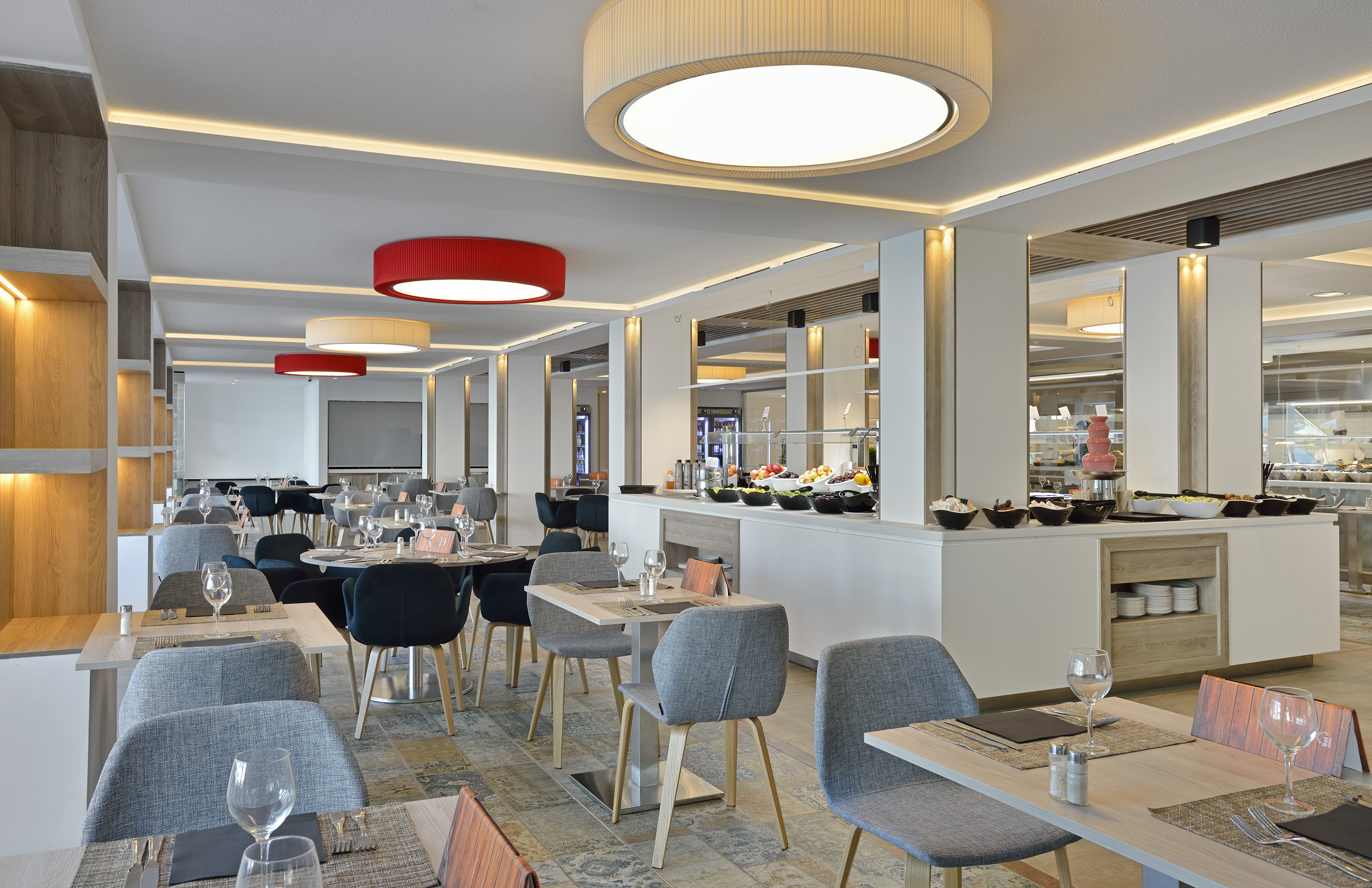 Modern hotel restaurant with buffet display, set dining tables, and gray chairs.
