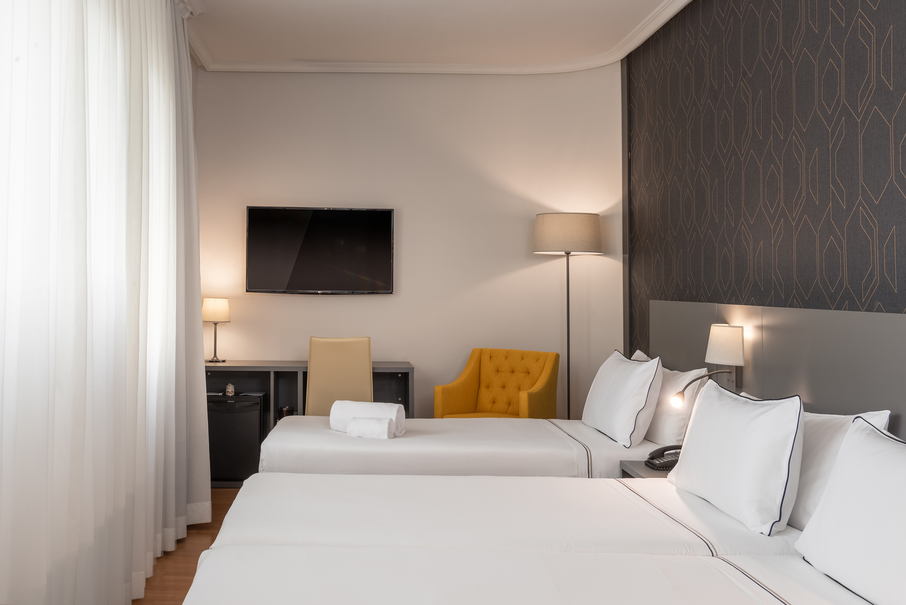 Hotel room with twin beds, TV, yellow armchair, and modern decor.