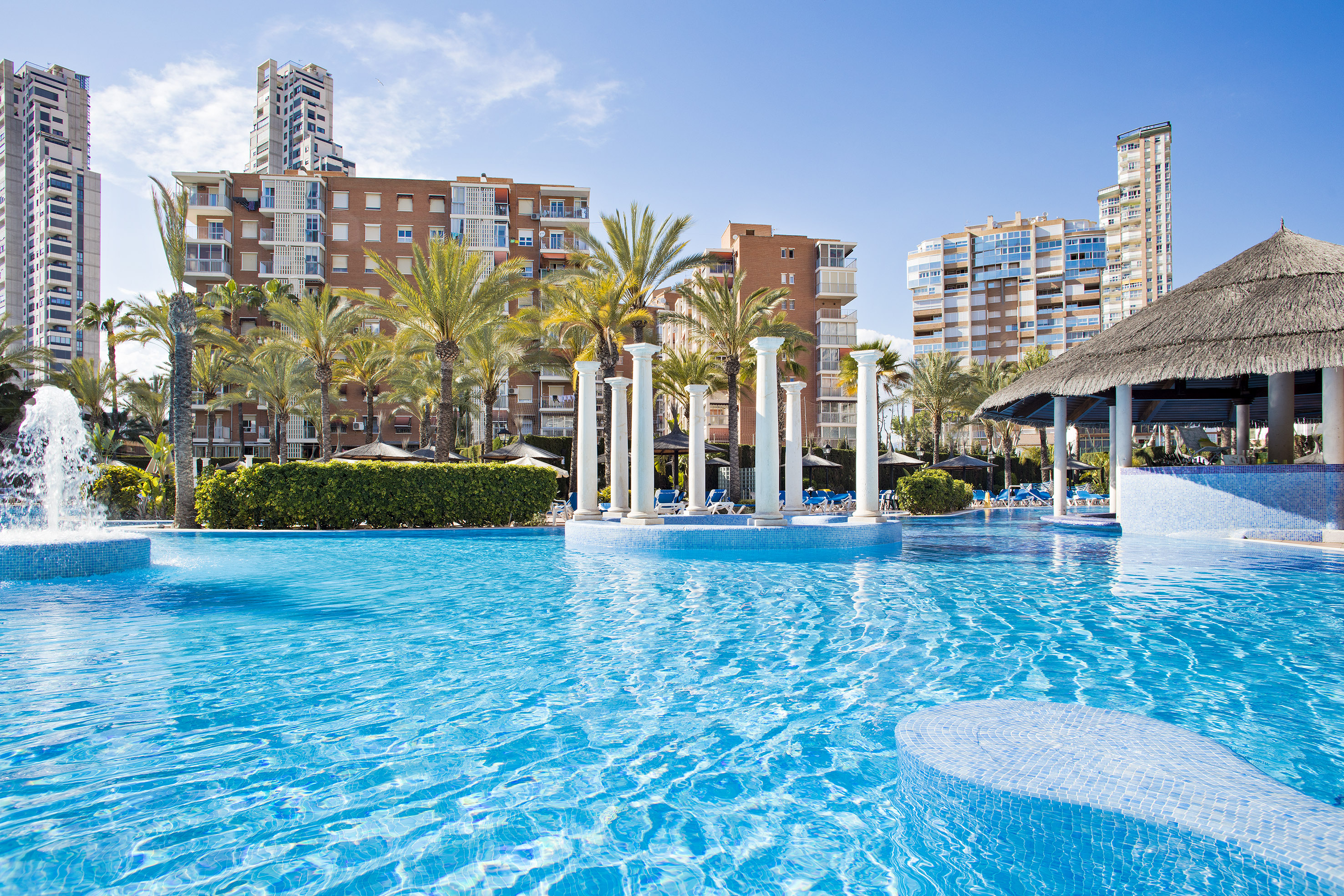Sunny resort pool with fountain, palm trees, columns, and modern hotel buildings.