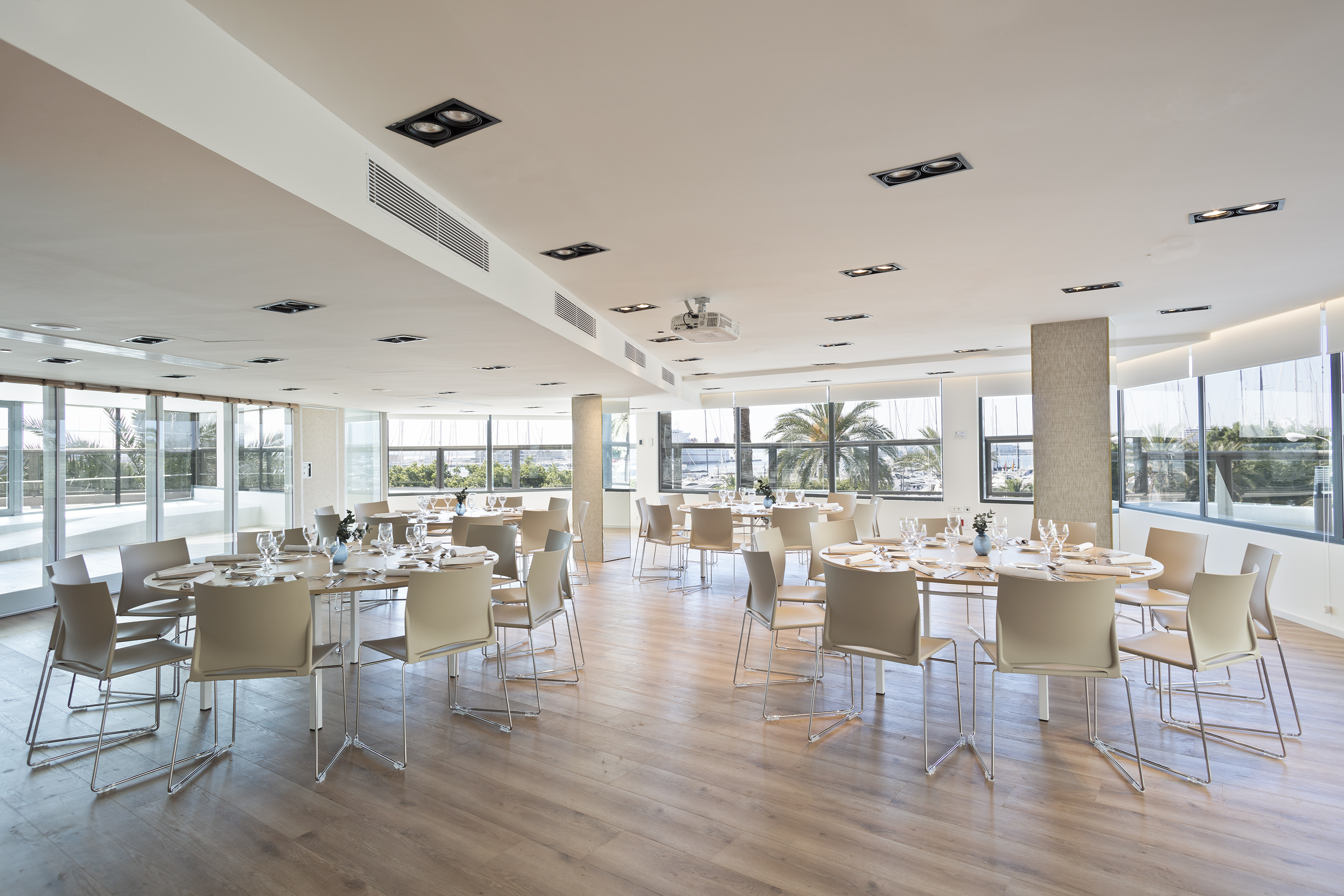 Modern event space with tables, beige chairs, wood floor, and marina view.