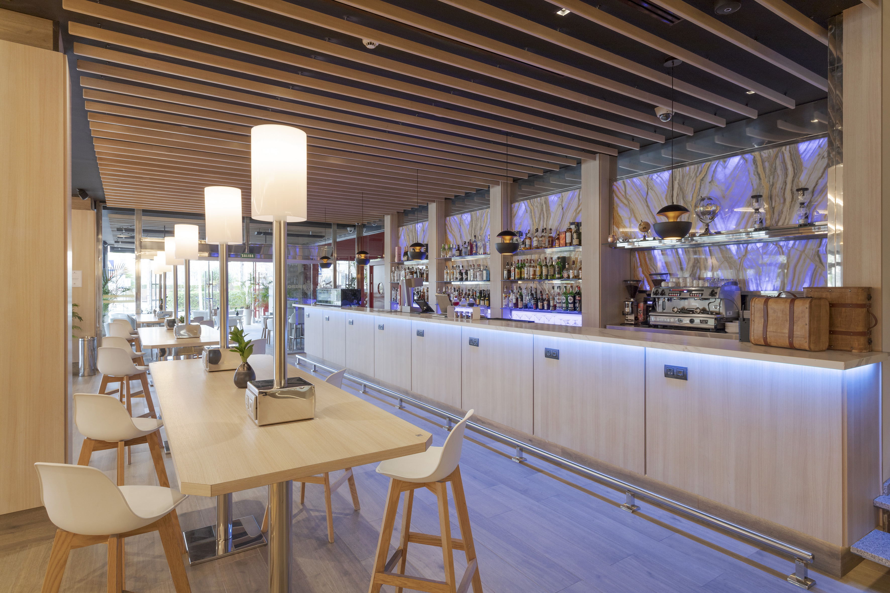 Modern bar and lounge with wooden counter, blue lighting, liquor, and dining tables.