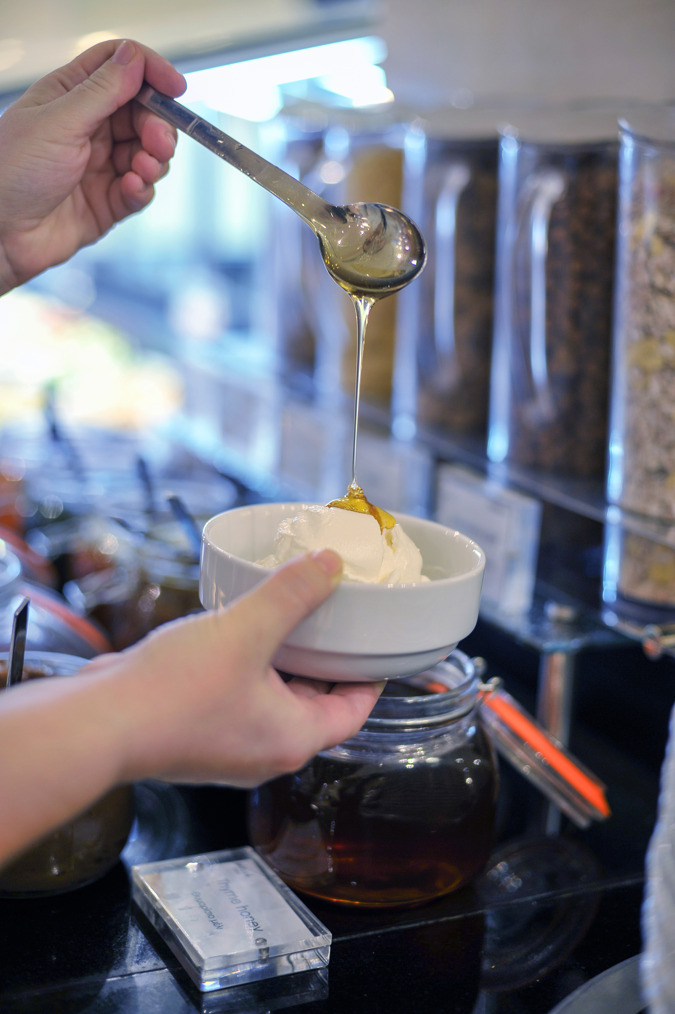Hand dripping thyme honey onto white yogurt in a bowl at a breakfast buffet.
