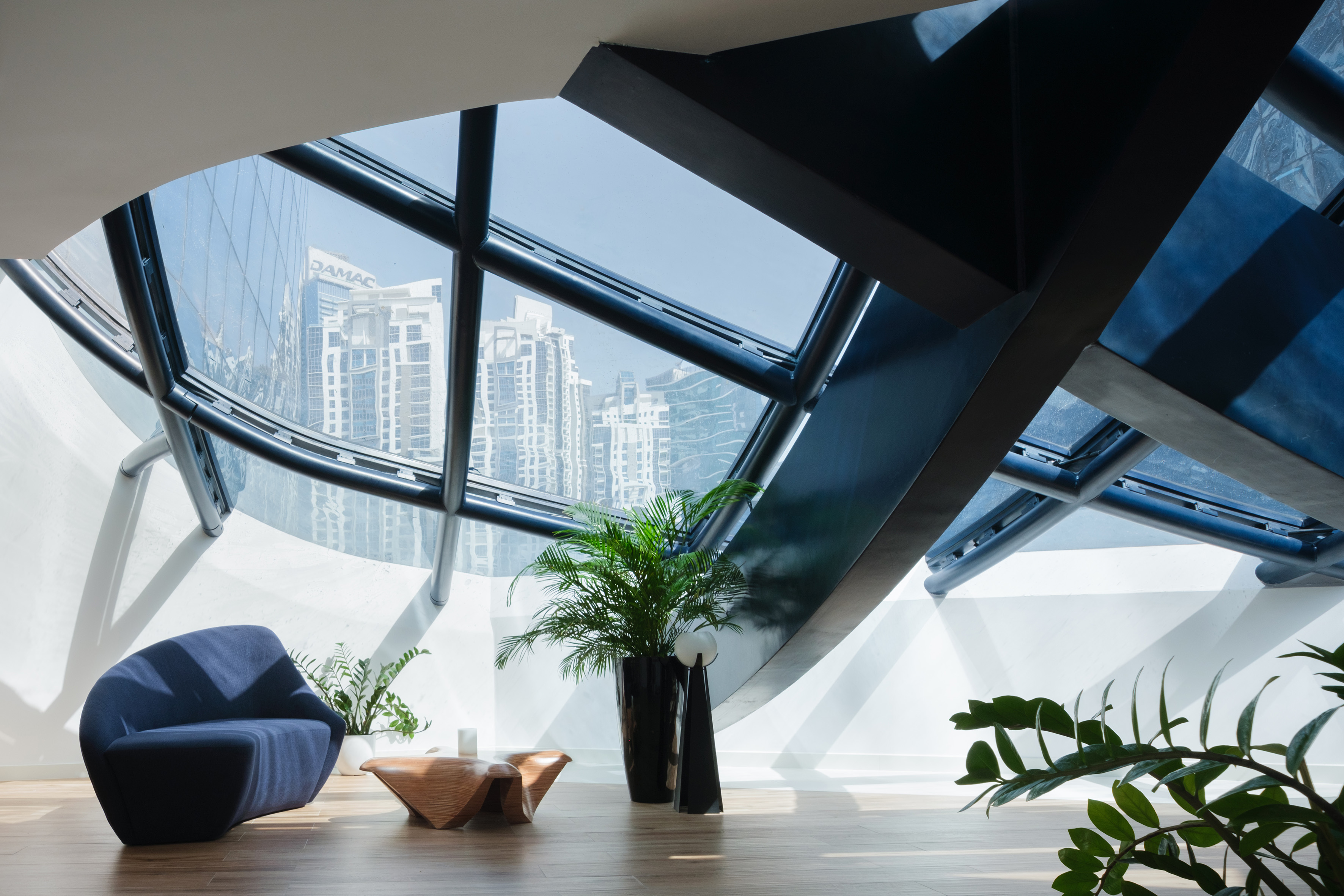 Modern lounge area with curved windows, city skyline, blue sofa, and green plants.