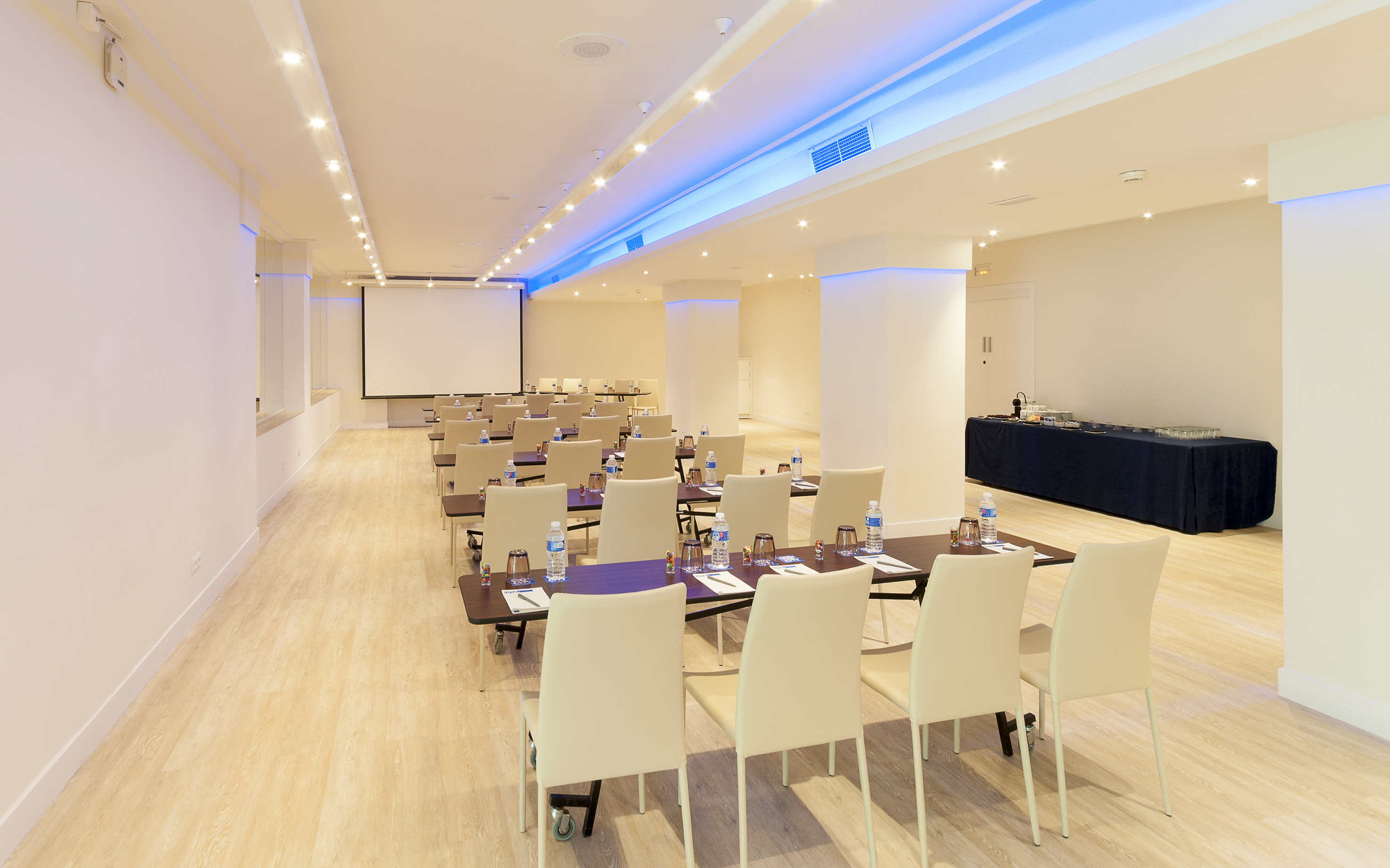 Bright modern conference room with screen, rows of tables, chairs, and blue lighting.