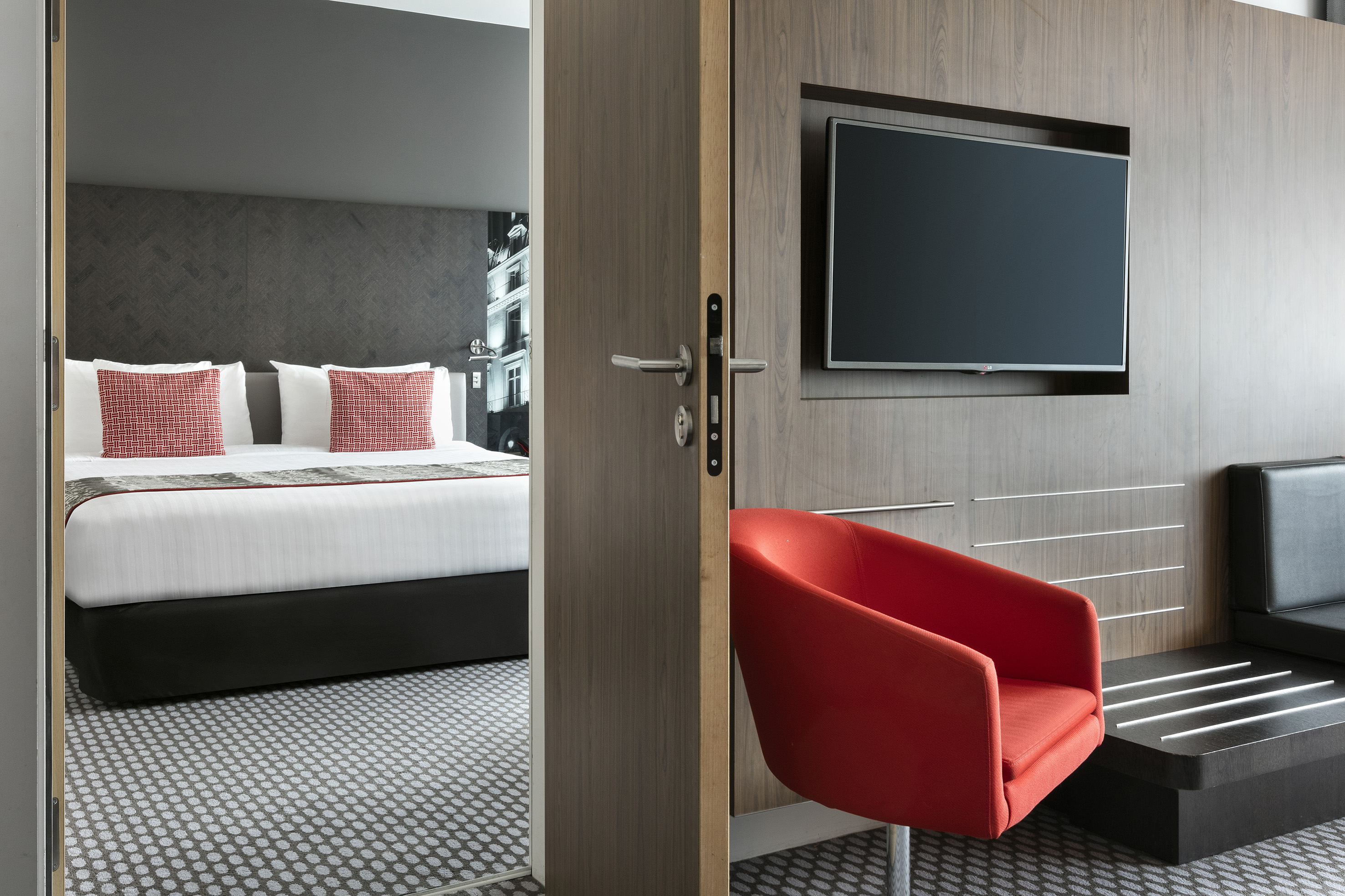 Modern hotel room: bedroom view, red armchair, wall TV, wood paneling.