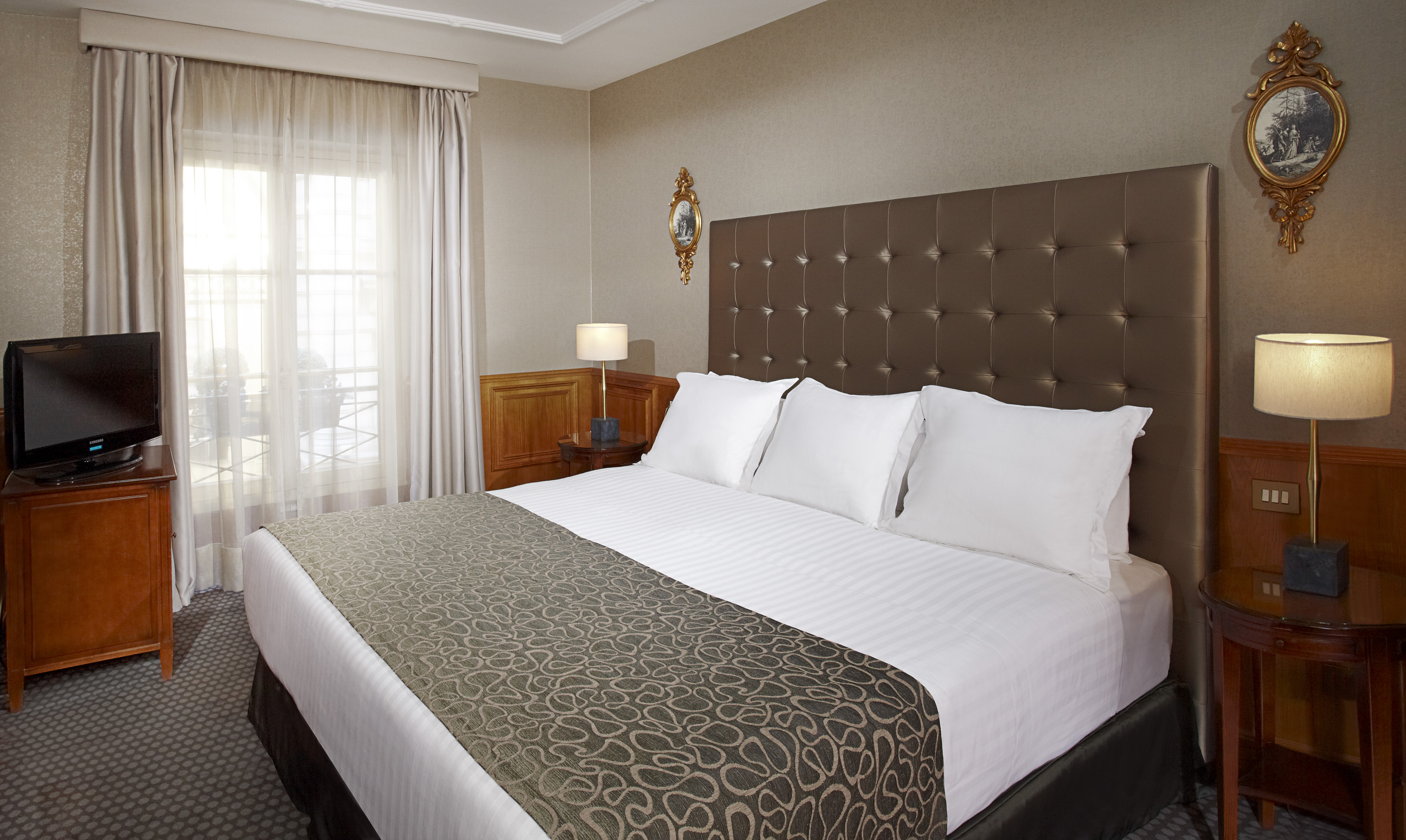 Hotel bedroom with king bed, brown headboard, white linens, and bright window.