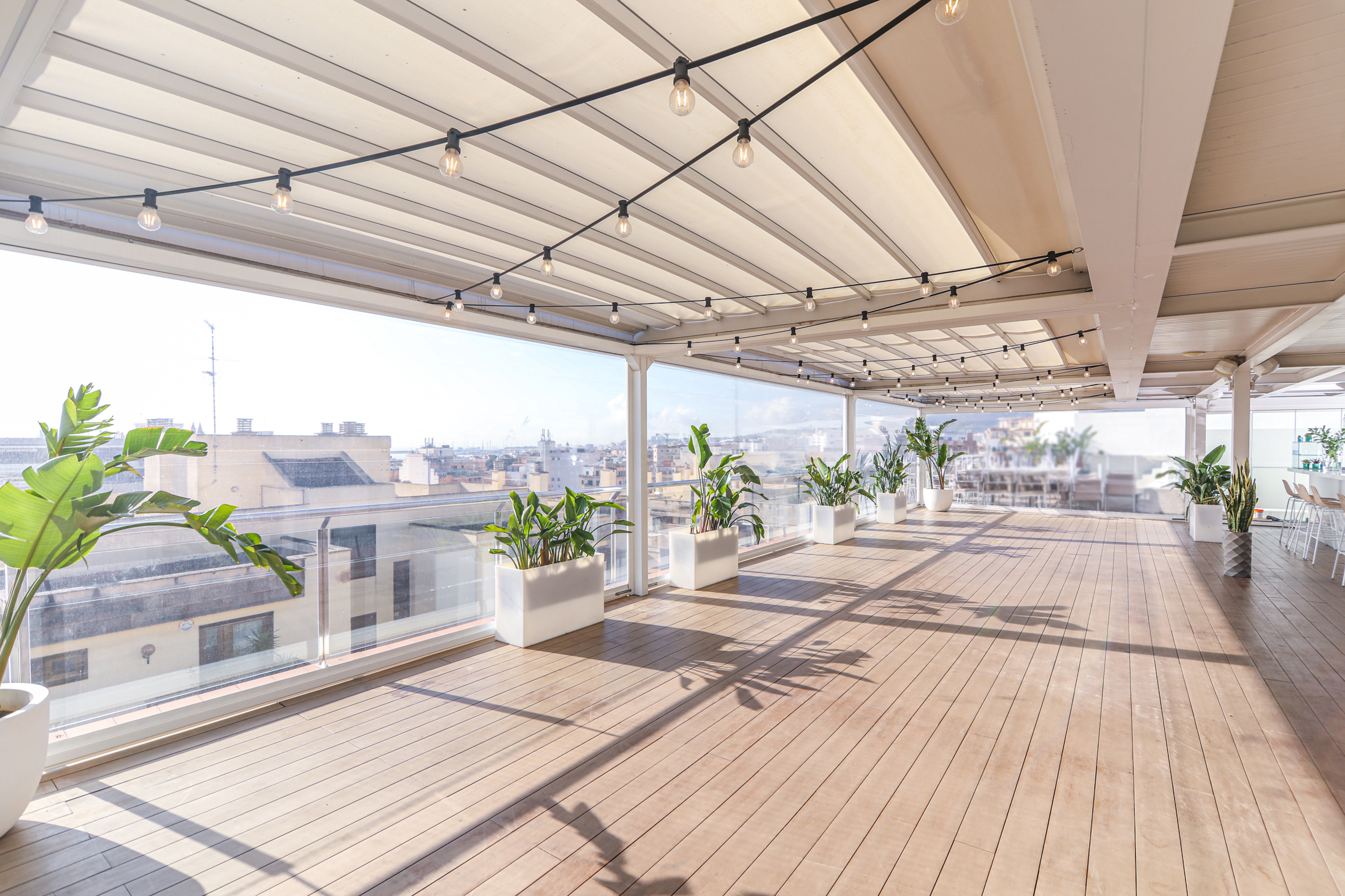 Rooftop terrace with wooden deck, string lights, potted plants, and city view.