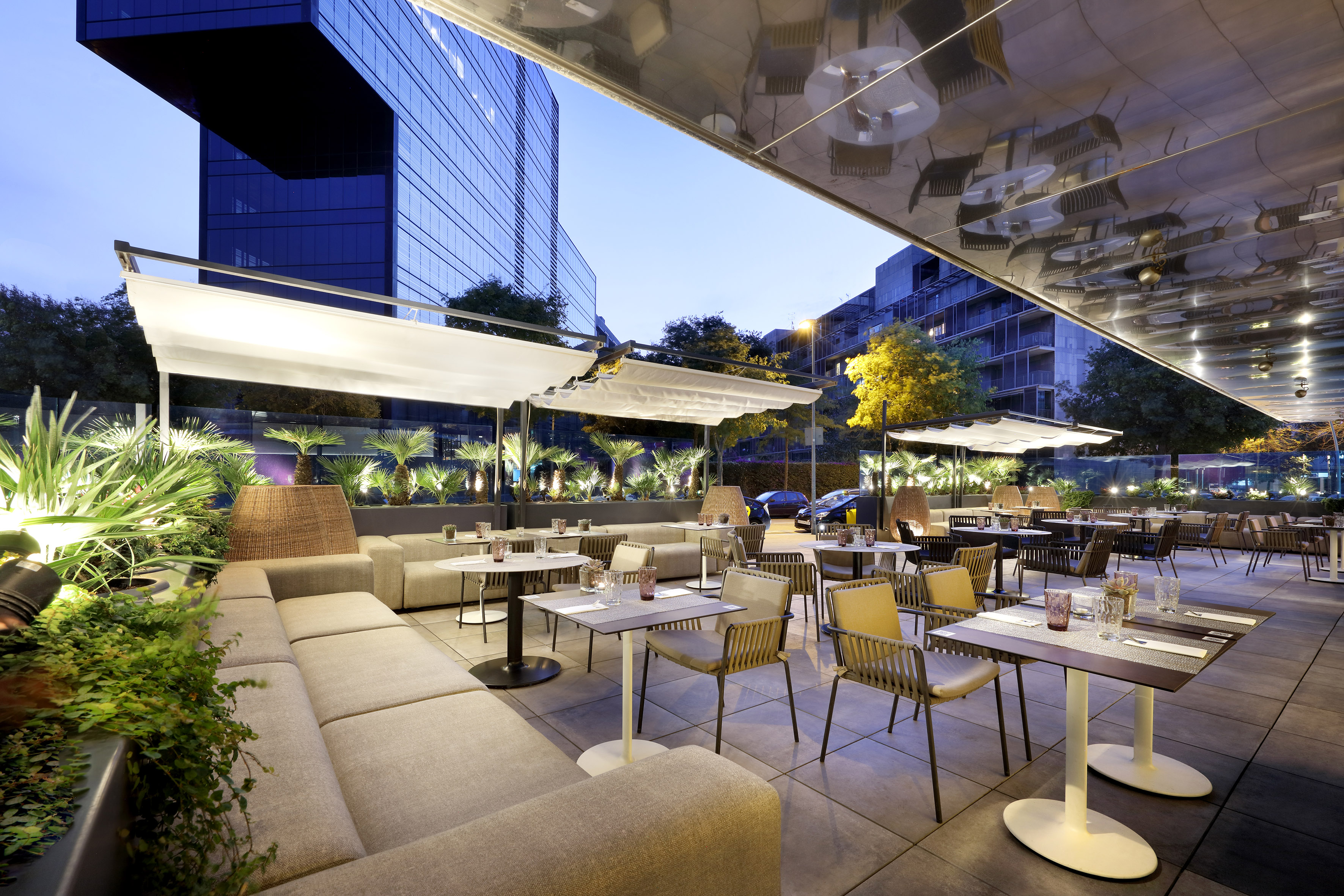 Modern urban restaurant terrace, outdoor dining, lounge seating, green plants.