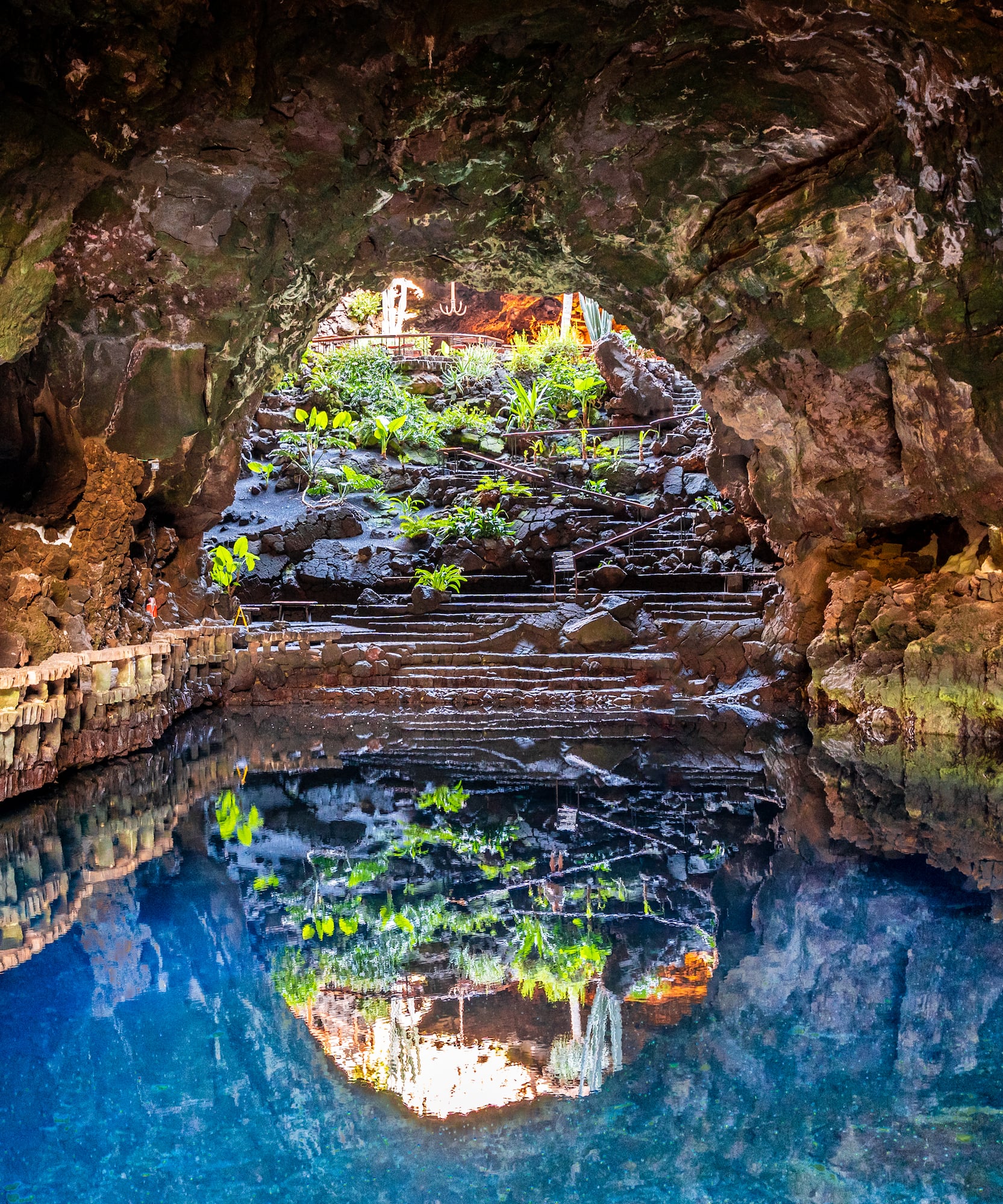 Jameos del Agua volcanic cave, blue lake reflecting entrance, plants, steps.