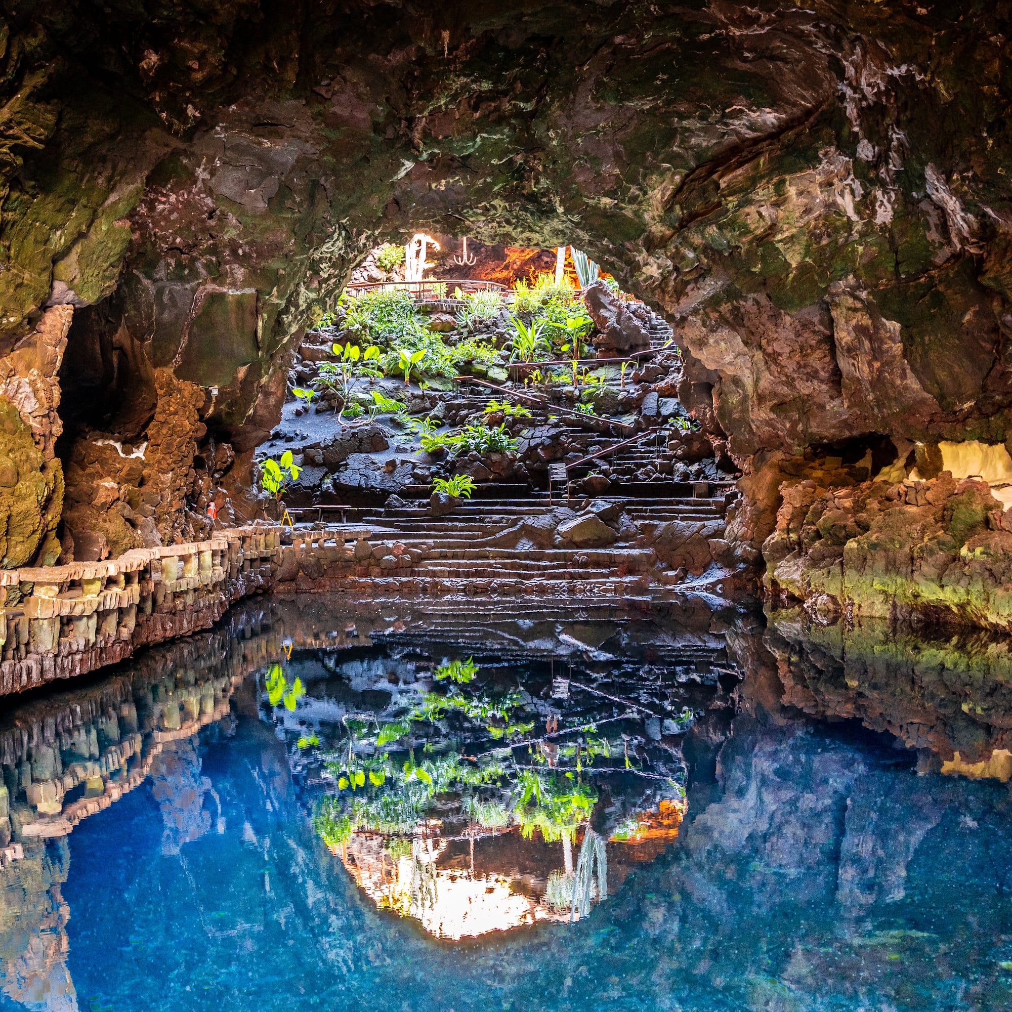 Jameos del Agua volcanic cave, blue lake reflecting entrance, plants, steps.