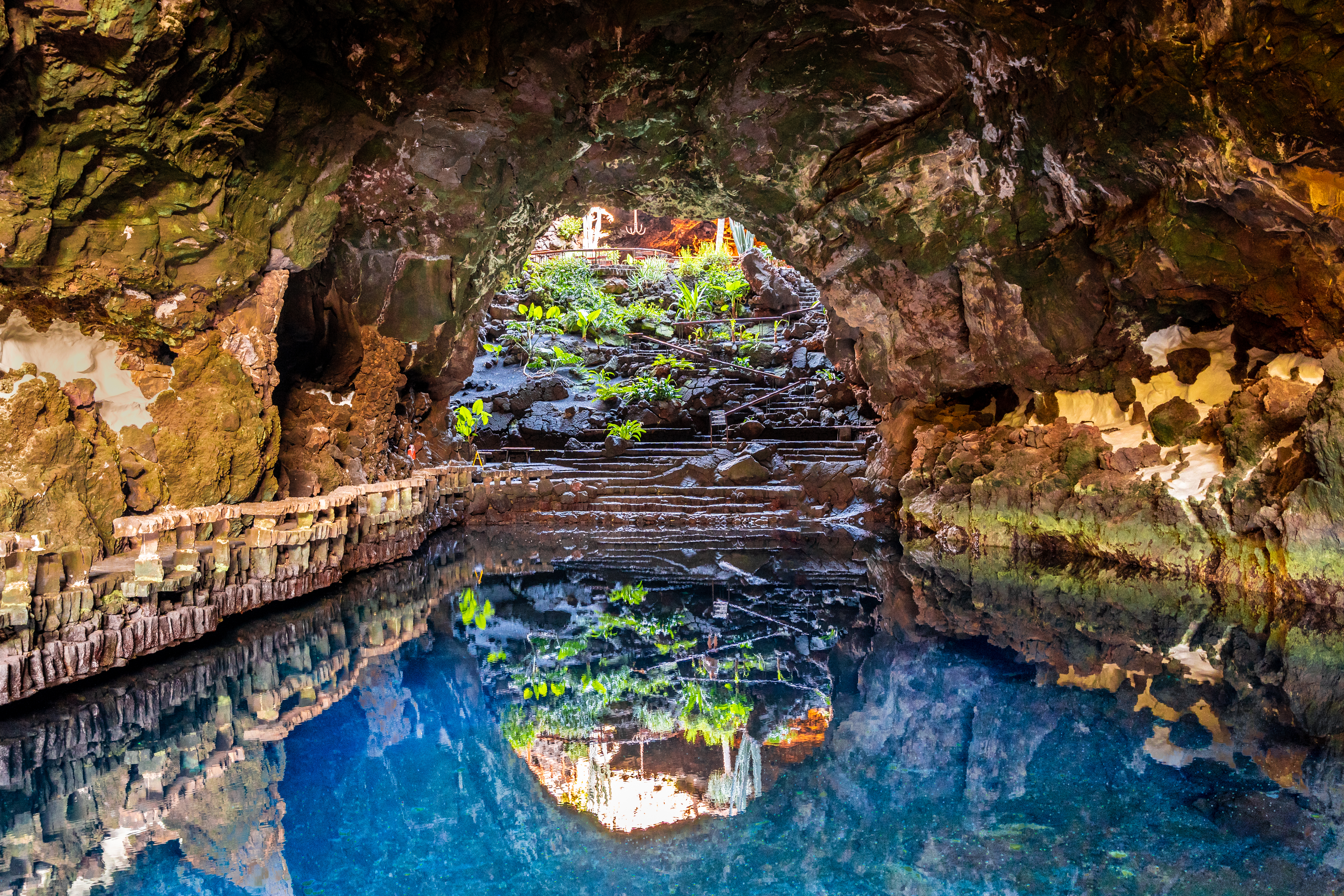 Jameos del Agua volcanic cave, blue lake reflecting entrance, plants, steps.