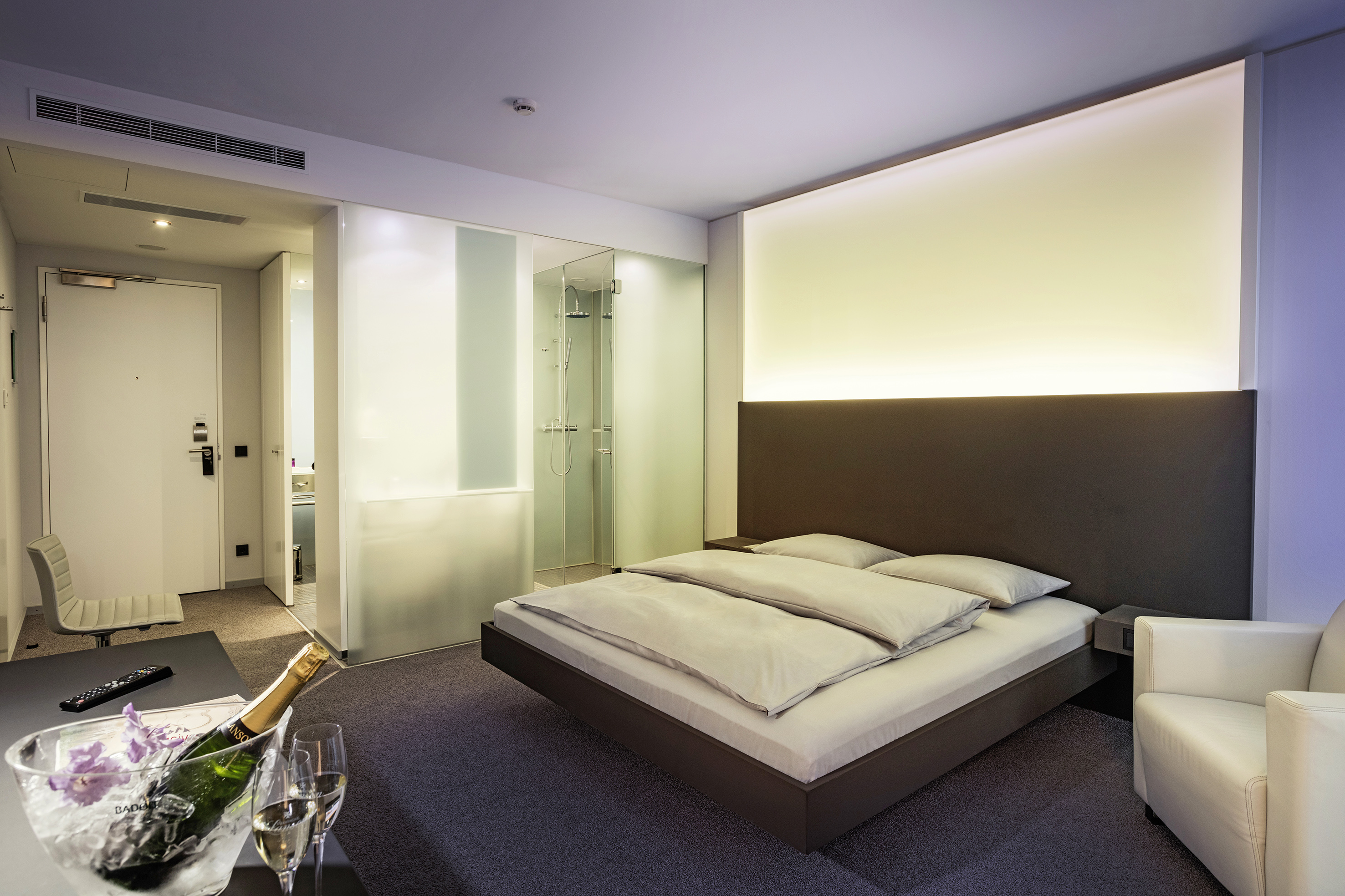 Modern hotel room, king bed, illuminated headboard, glass shower, champagne.