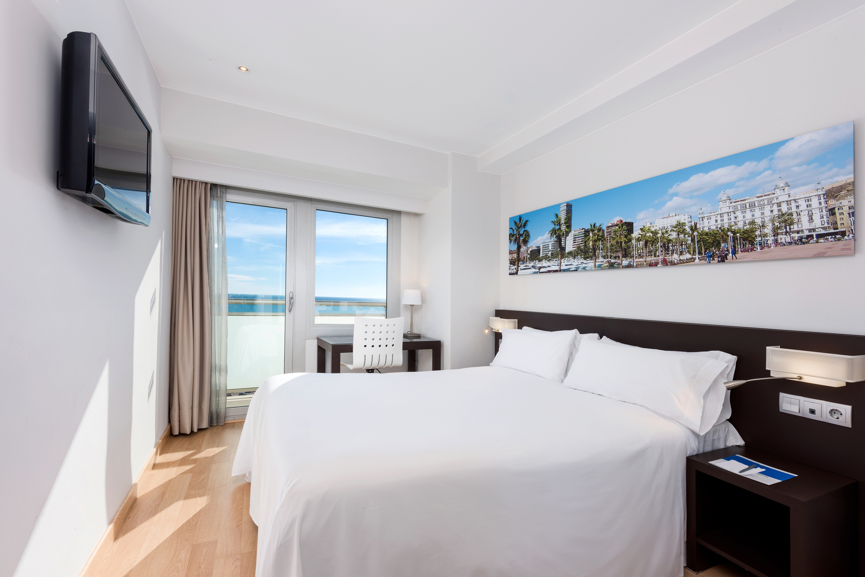 Modern hotel room with king bed, sea view balcony, desk, and panoramic city art.