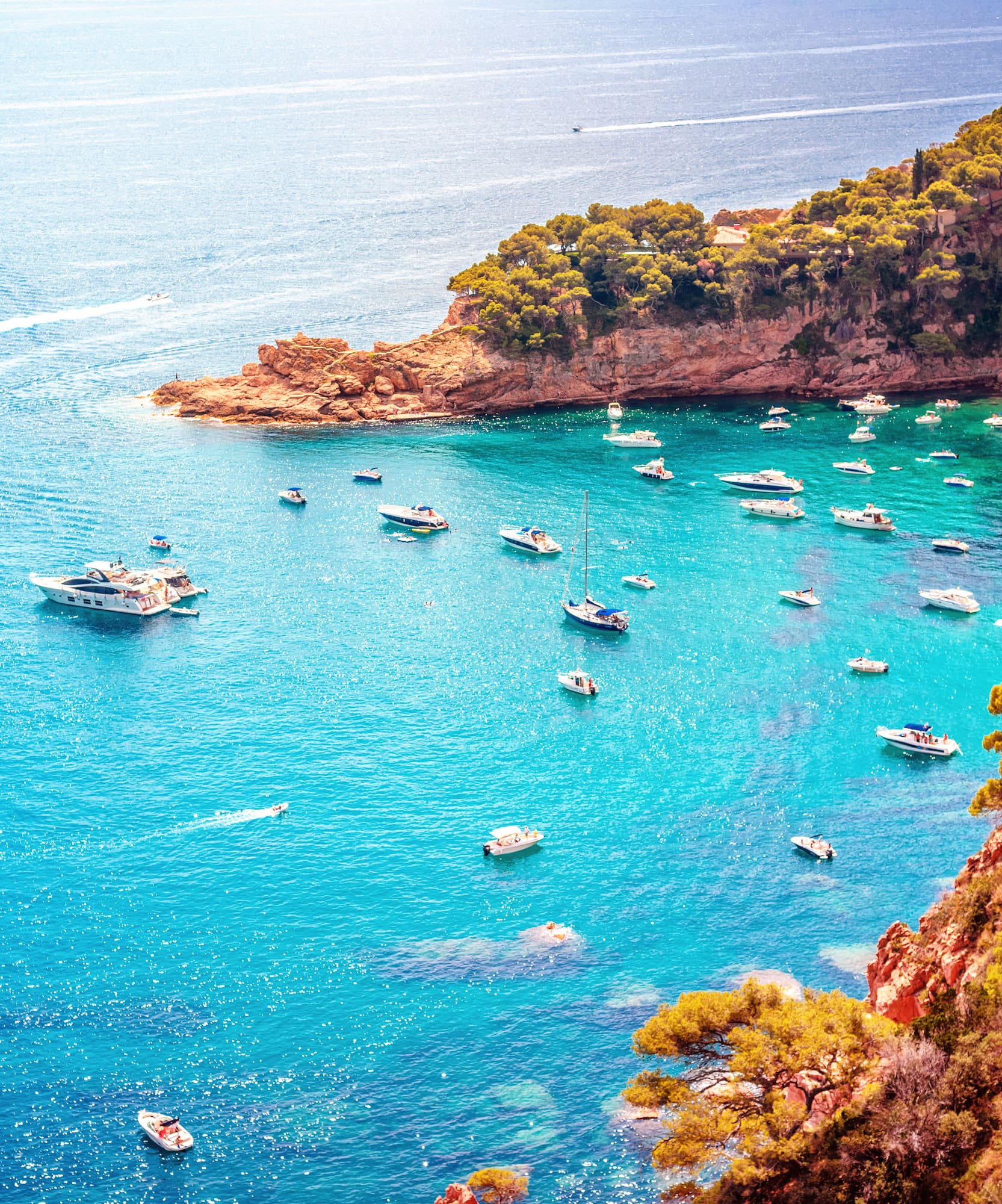 Boats on clear turquoise Mediterranean bay, rocky coast, green trees.