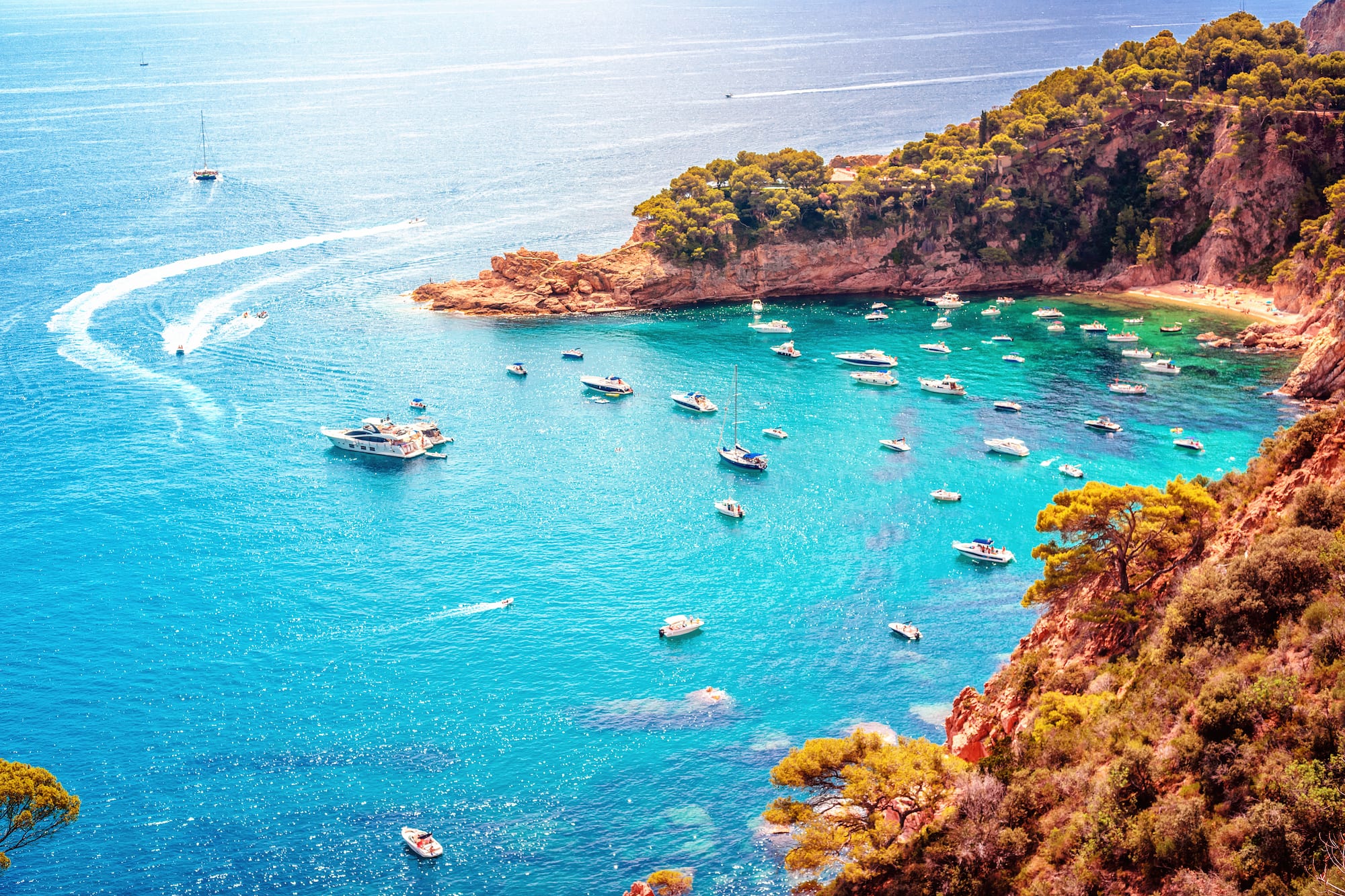 Boats on clear turquoise Mediterranean bay, rocky coast, green trees.