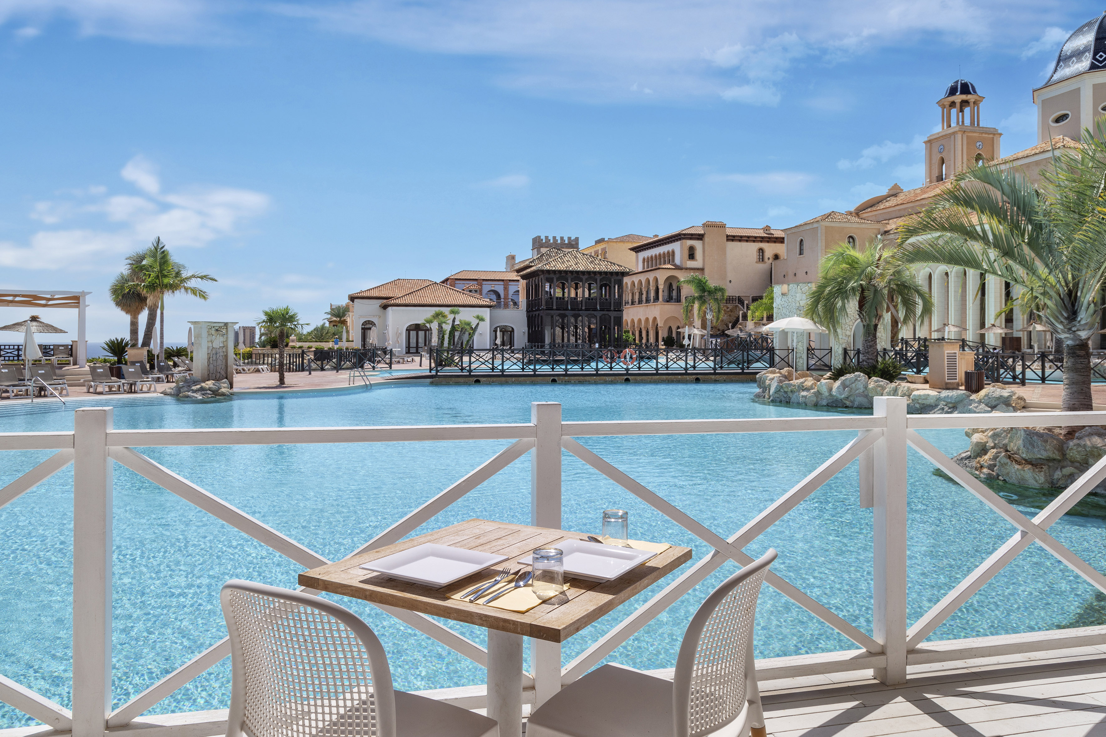 Dining table at a Mediterranean resort with a large blue swimming pool.