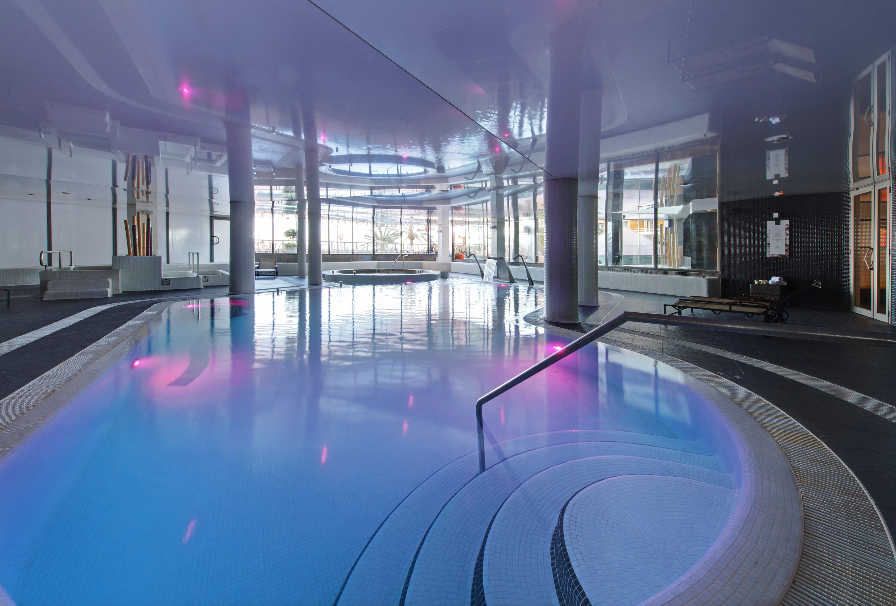 Indoor spa swimming pool with blue illuminated water, jacuzzi, and large windows.
