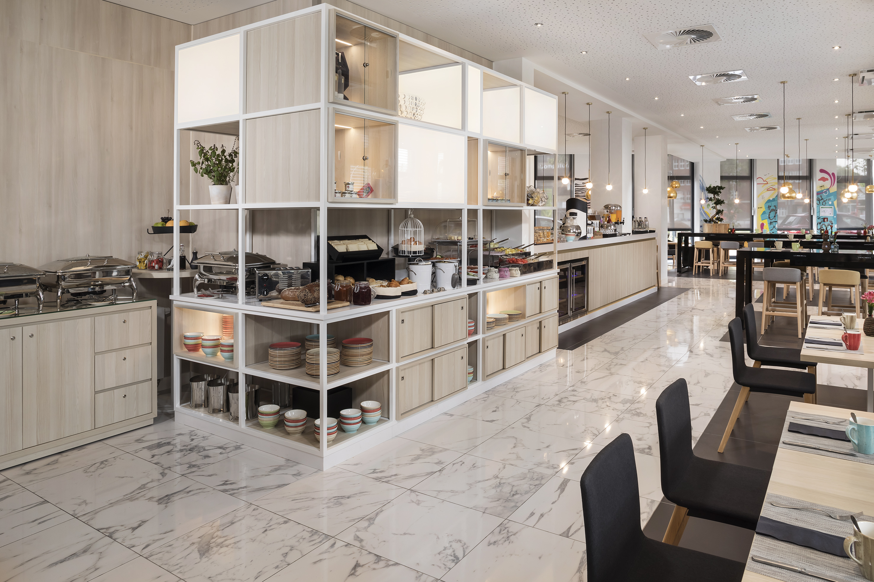 Bright hotel breakfast room with buffet counter, food displays, and guest seating.