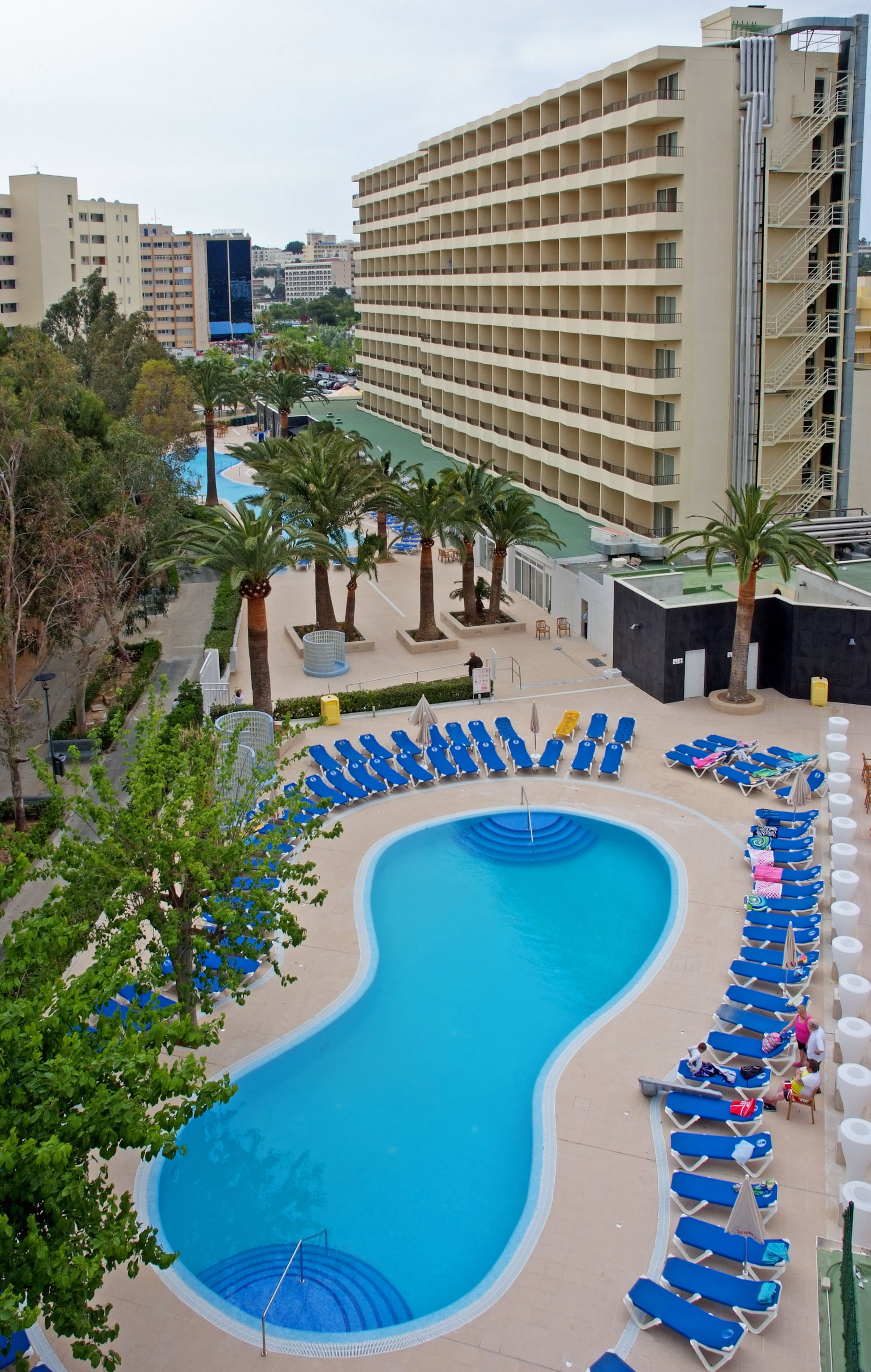 Resort pool deck with blue sun loungers, palm trees, and hotel building.