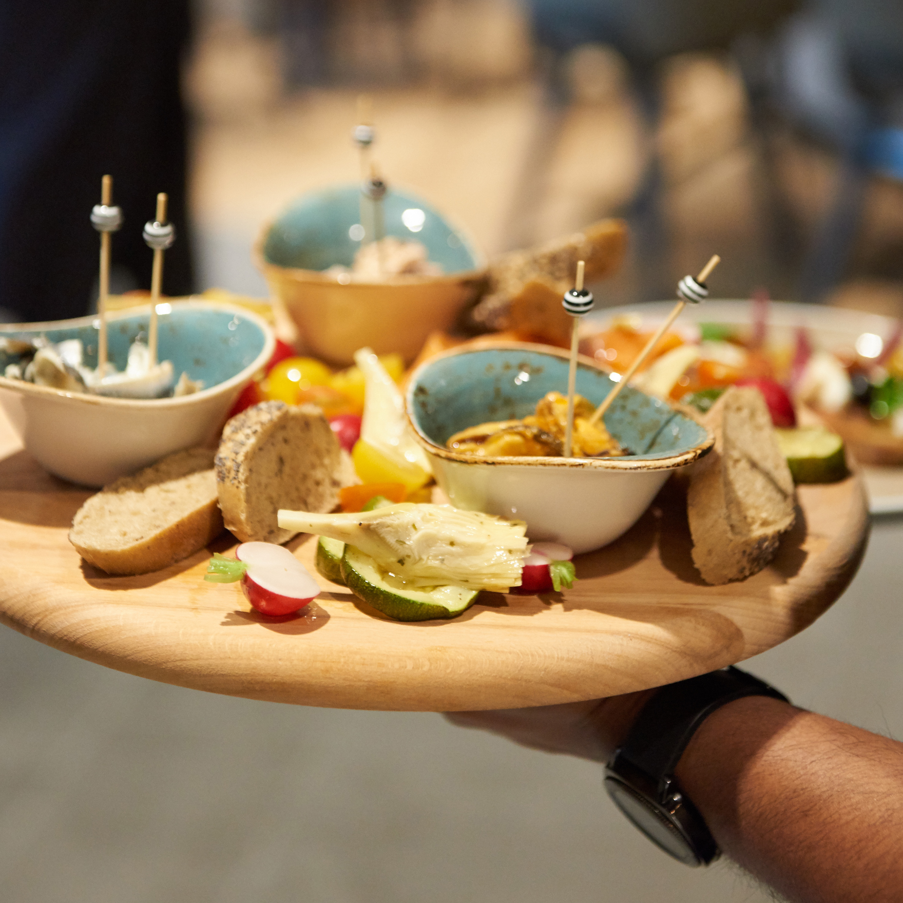 Assorted appetizers on a wooden board: small bowls, bread, artichoke, radishes.