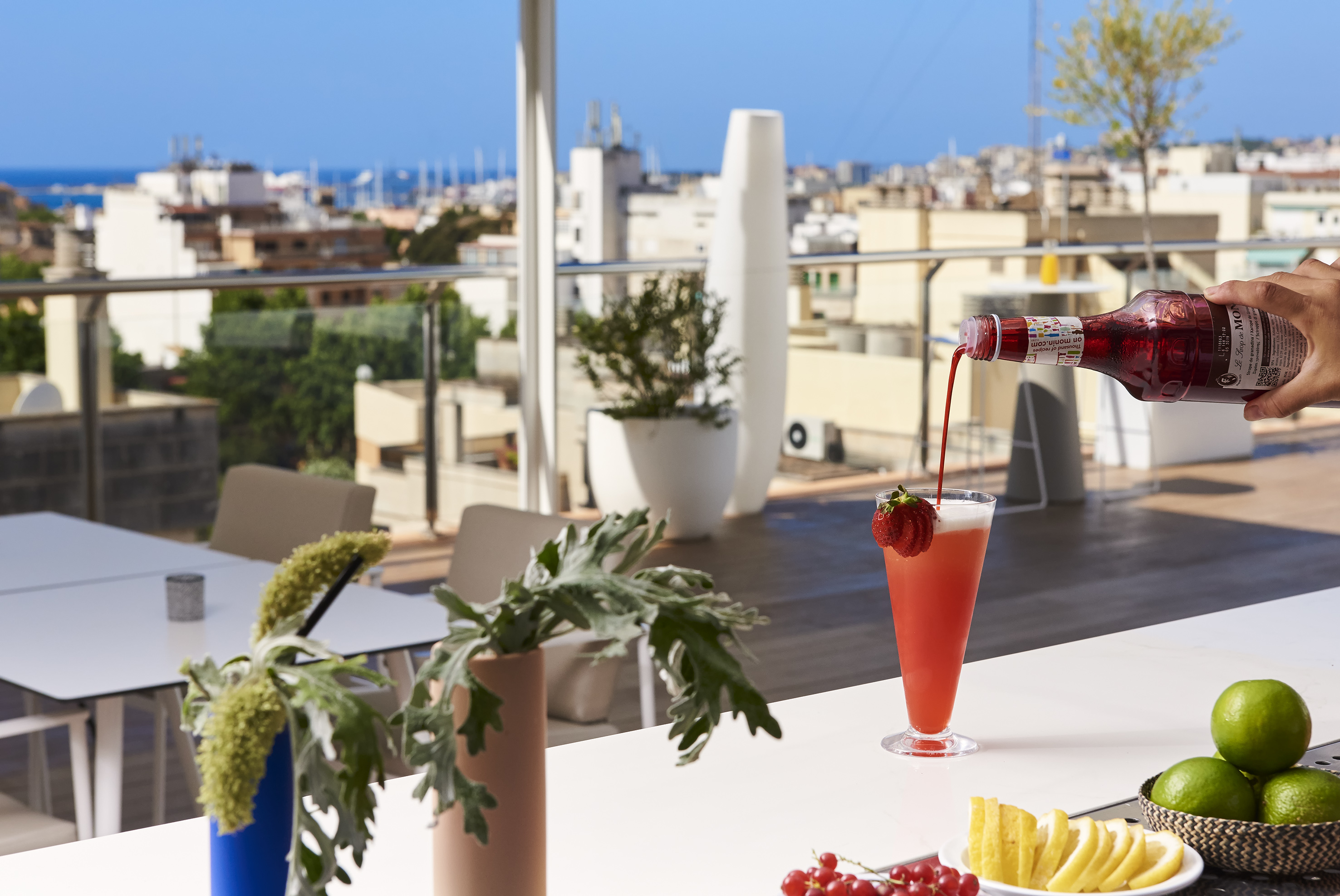 Pouring Monin syrup into a strawberry cocktail on a sunny rooftop bar with city view.