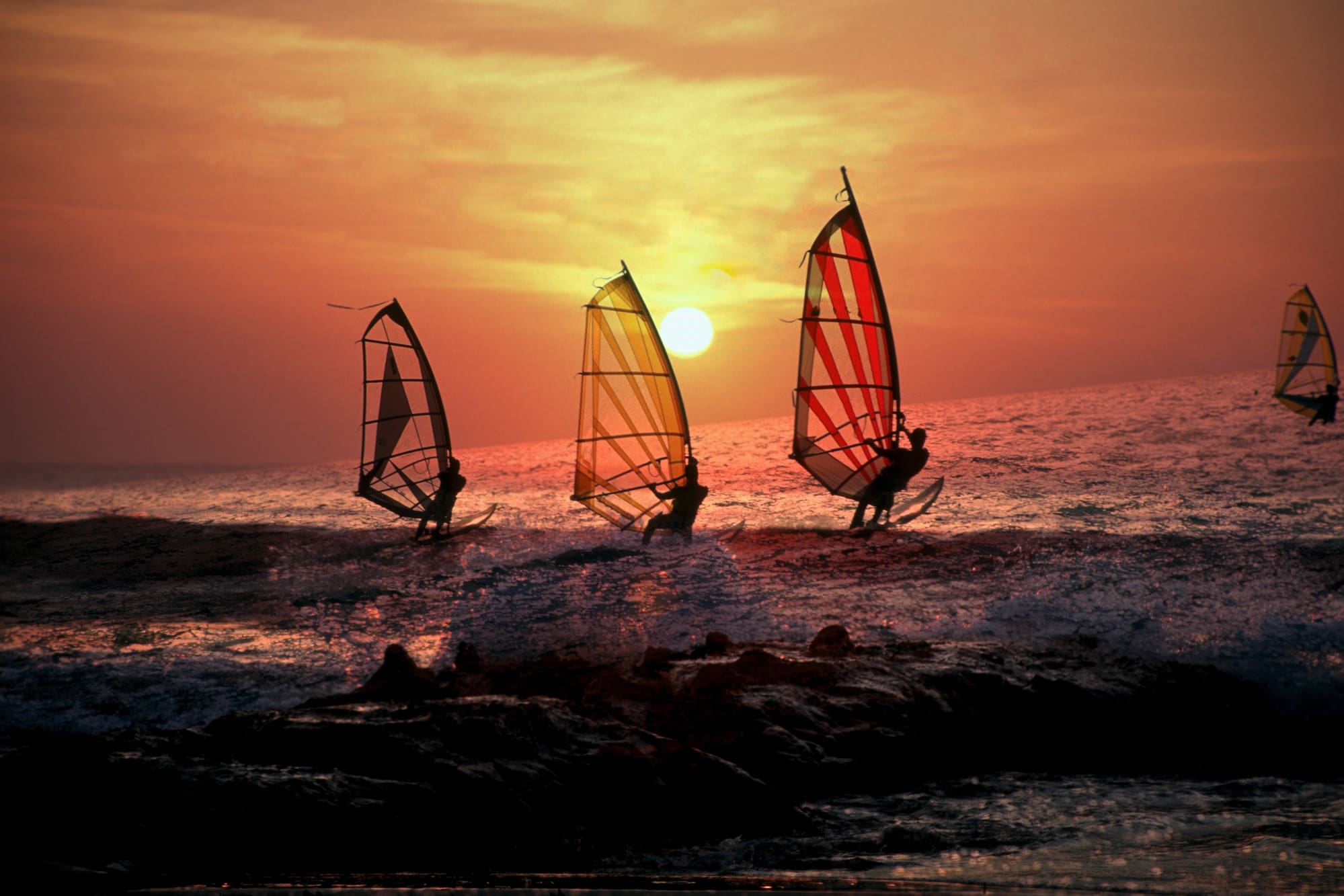 Windsurfistas en el mar al atardecer, siluetas con velas coloridas bajo el sol.