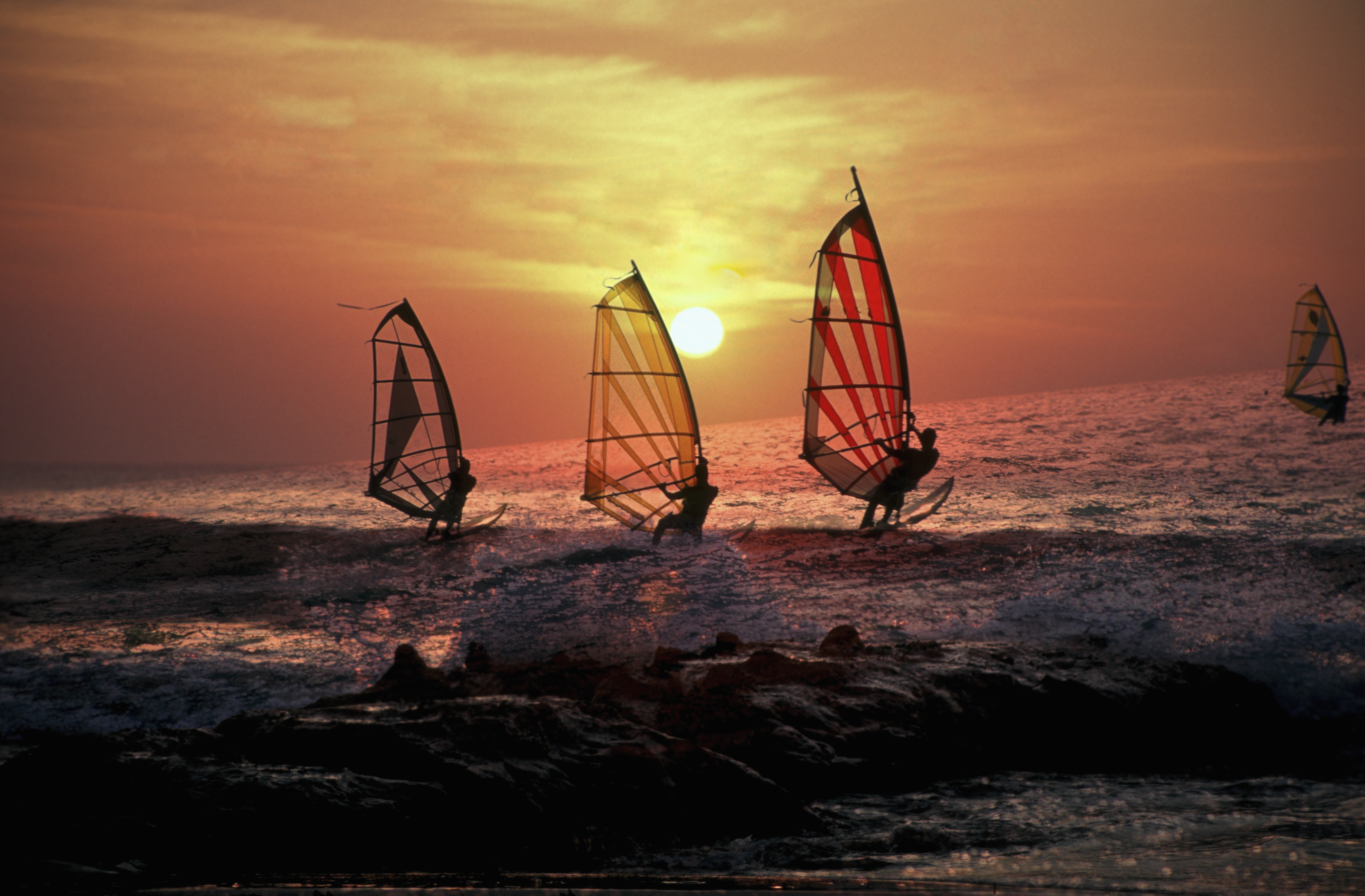 Windsurfistas en el mar al atardecer, siluetas con velas coloridas bajo el sol.