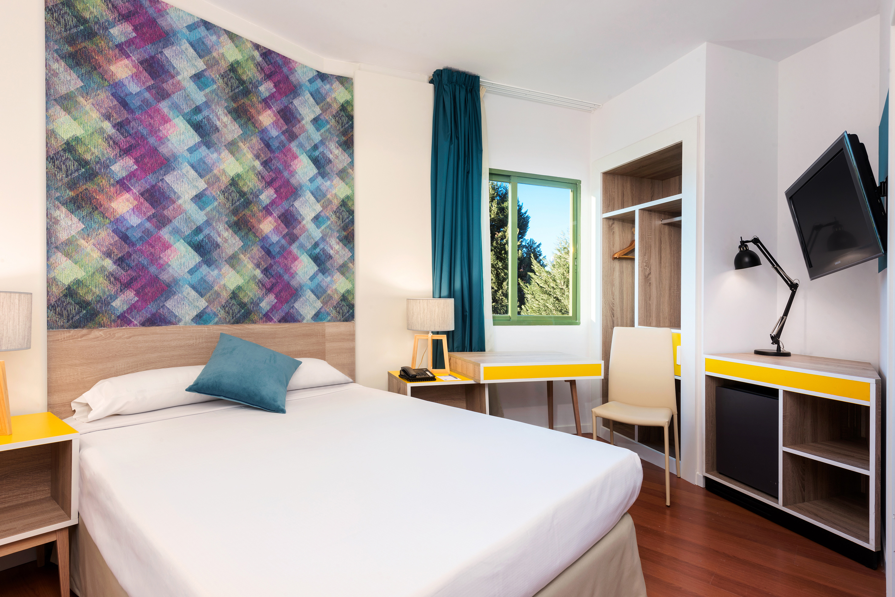 Modern hotel bedroom with vibrant geometric wallpaper, bed, desk, and TV.
