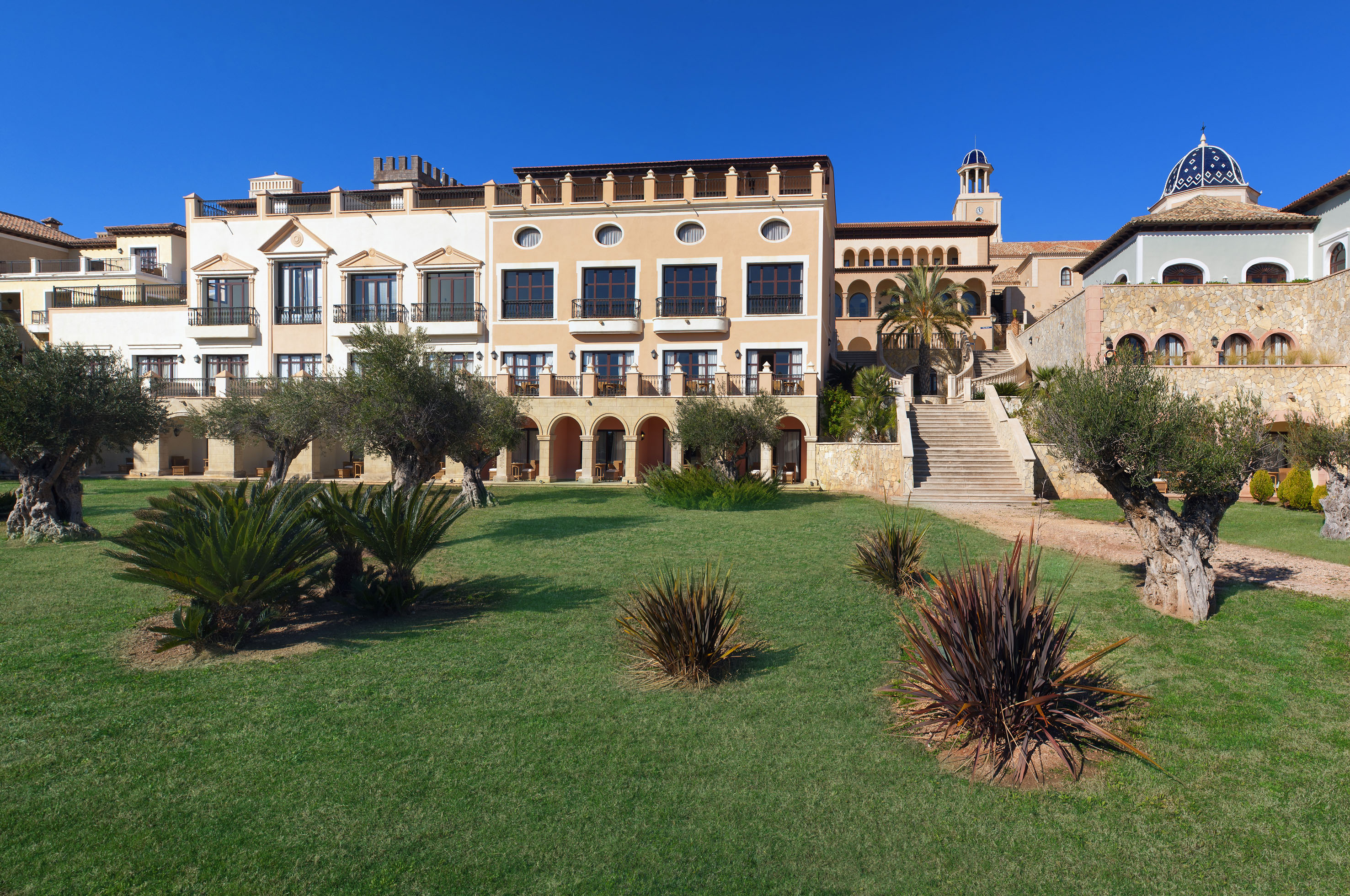 Sunny Mediterranean resort hotel, green garden, olive trees, and tiled dome.