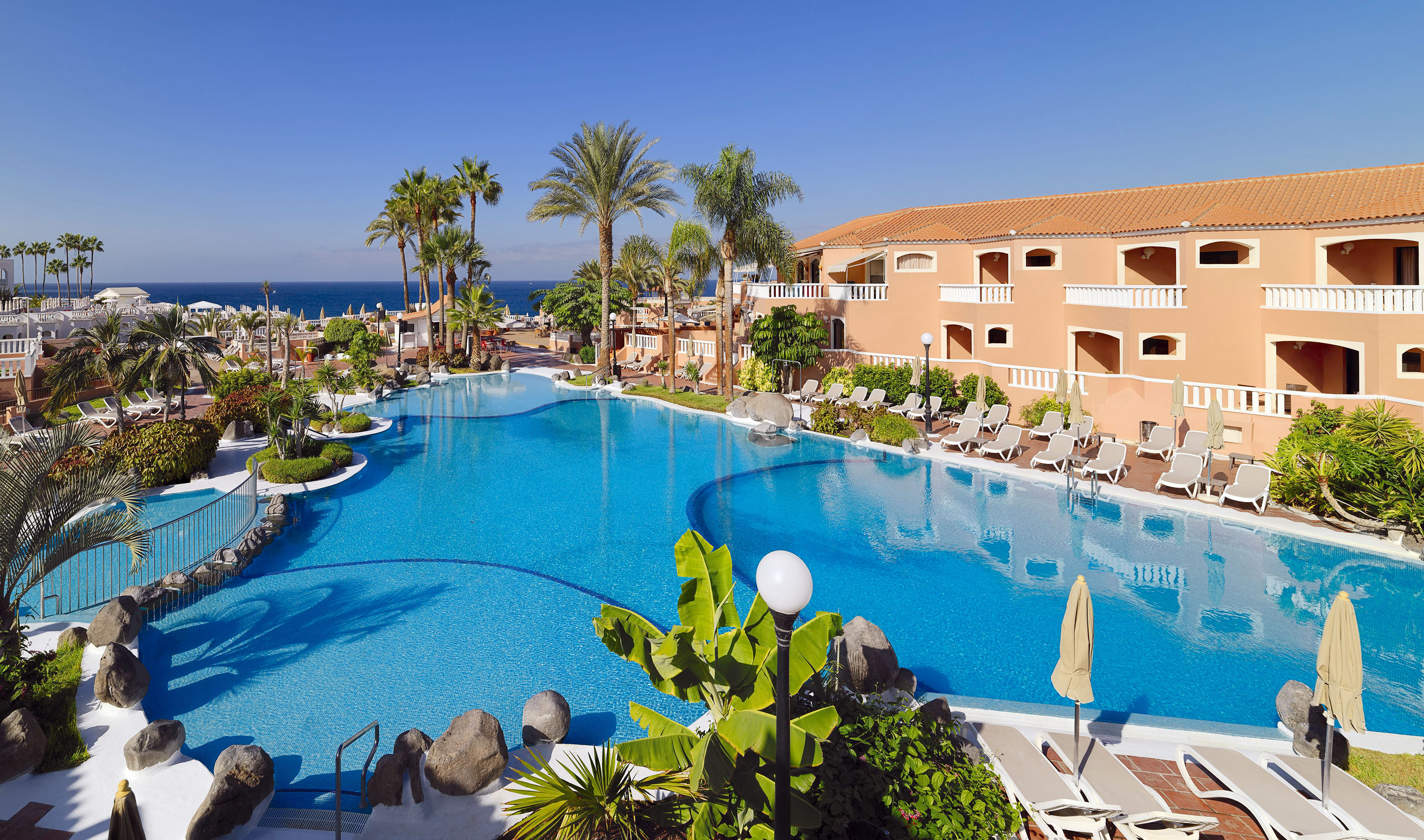 Hotel resort pool with palm trees, sun loungers, and ocean view.