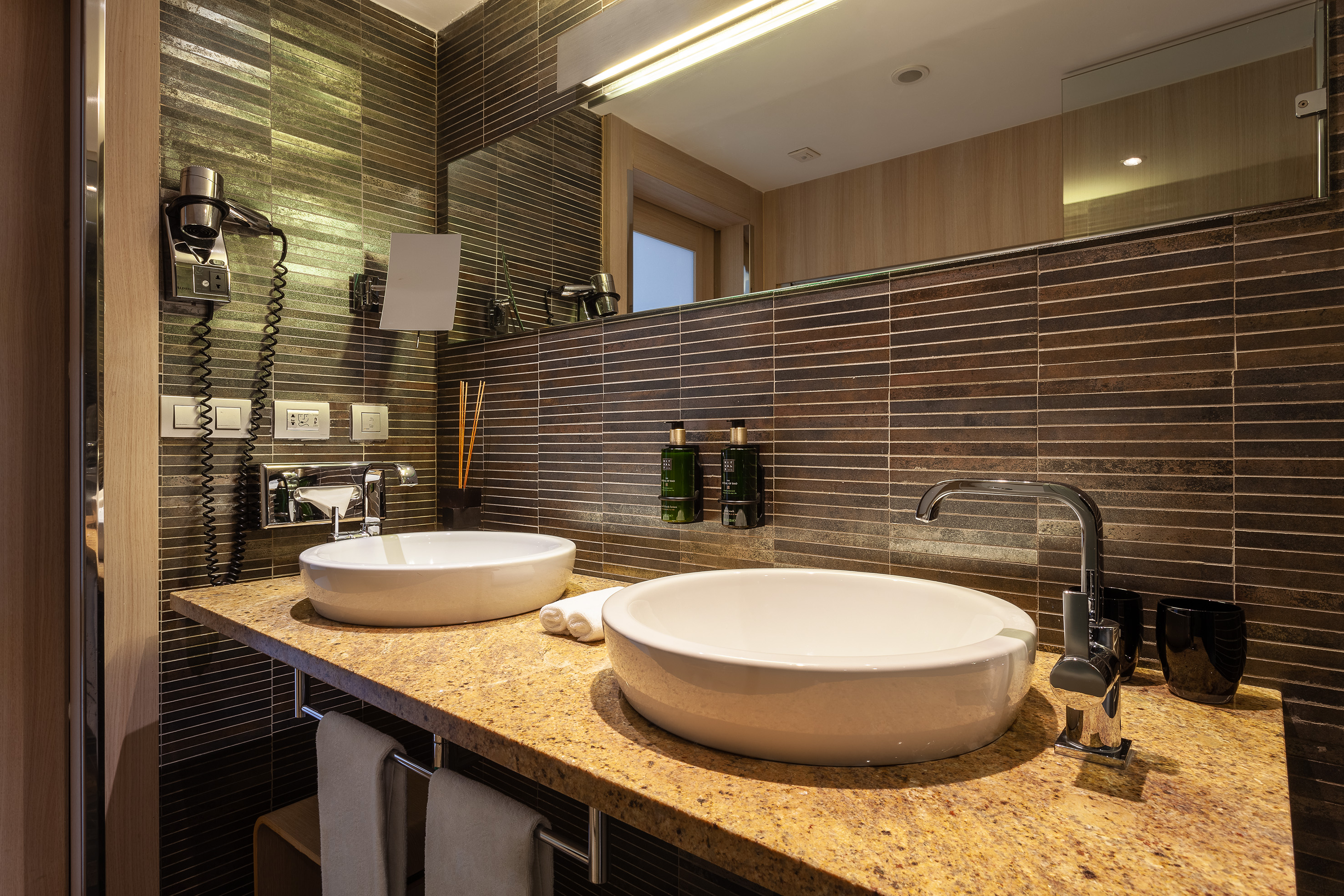 Modern hotel bathroom with double vessel sinks, granite counter, and tiled wall.