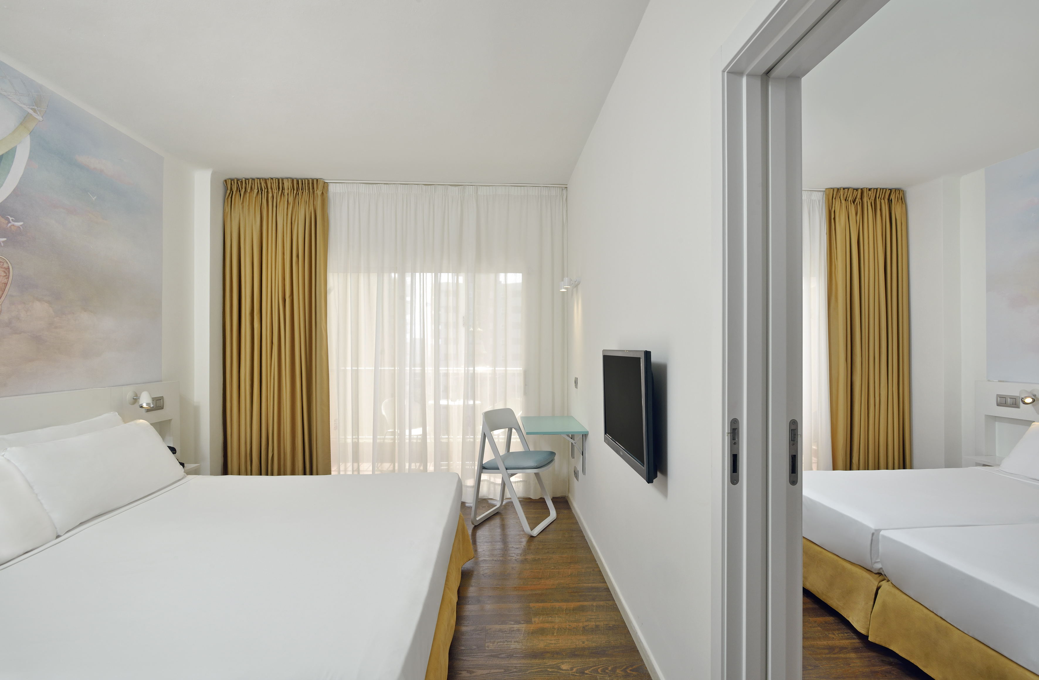 Modern hotel room with white bed, wooden floor, mustard curtains, desk, and TV.