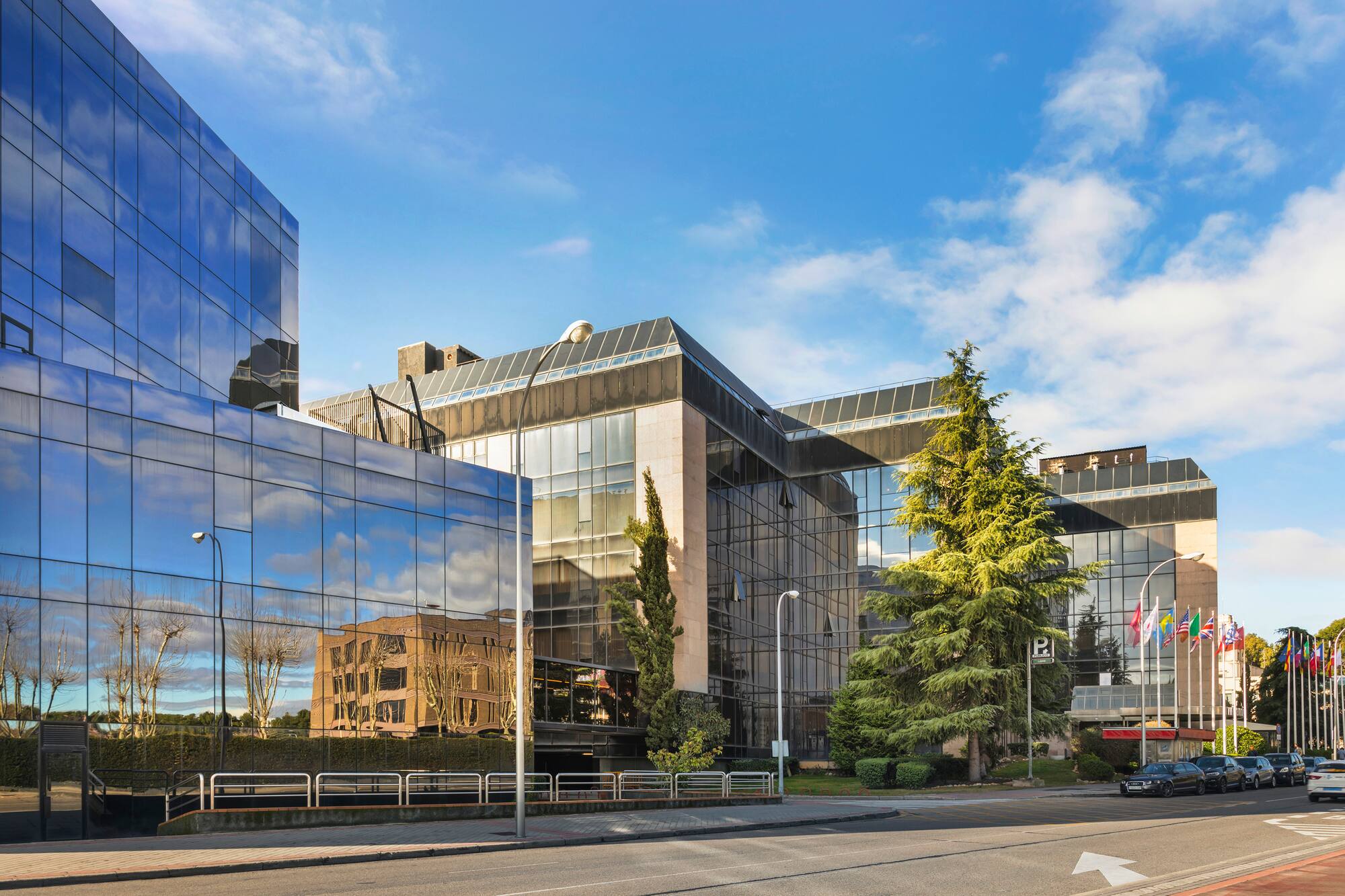 Modern reflective glass office building with corporate flags and street parking.