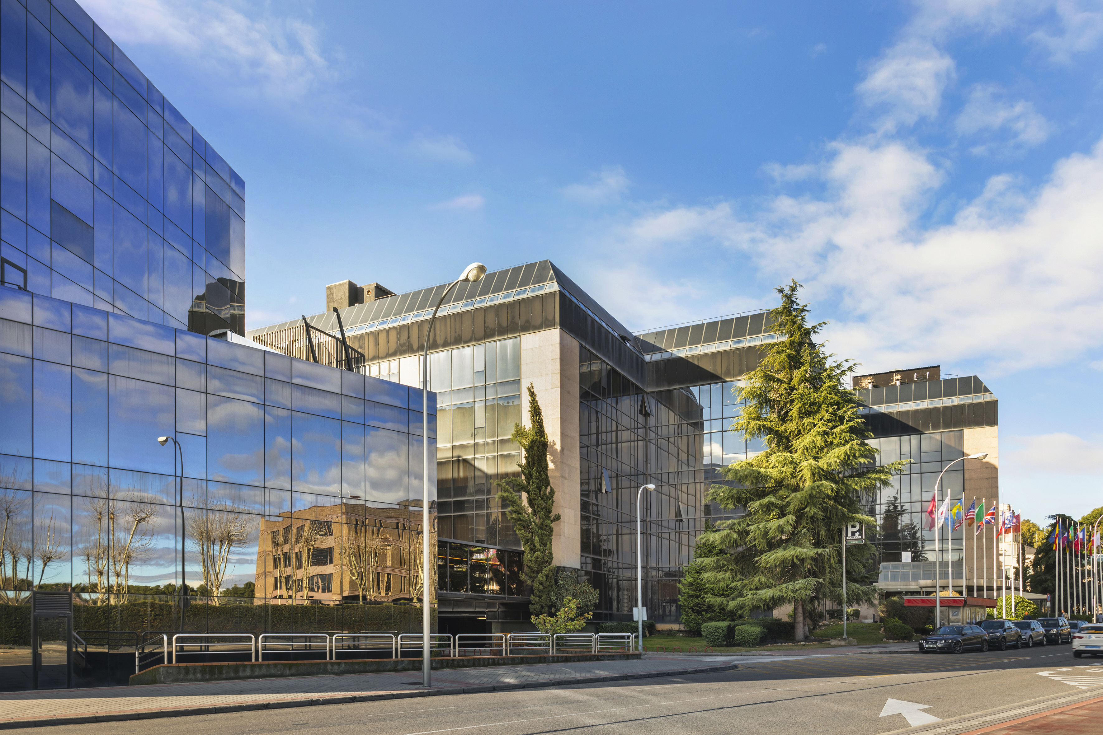 Modern reflective glass office building with corporate flags and street parking.