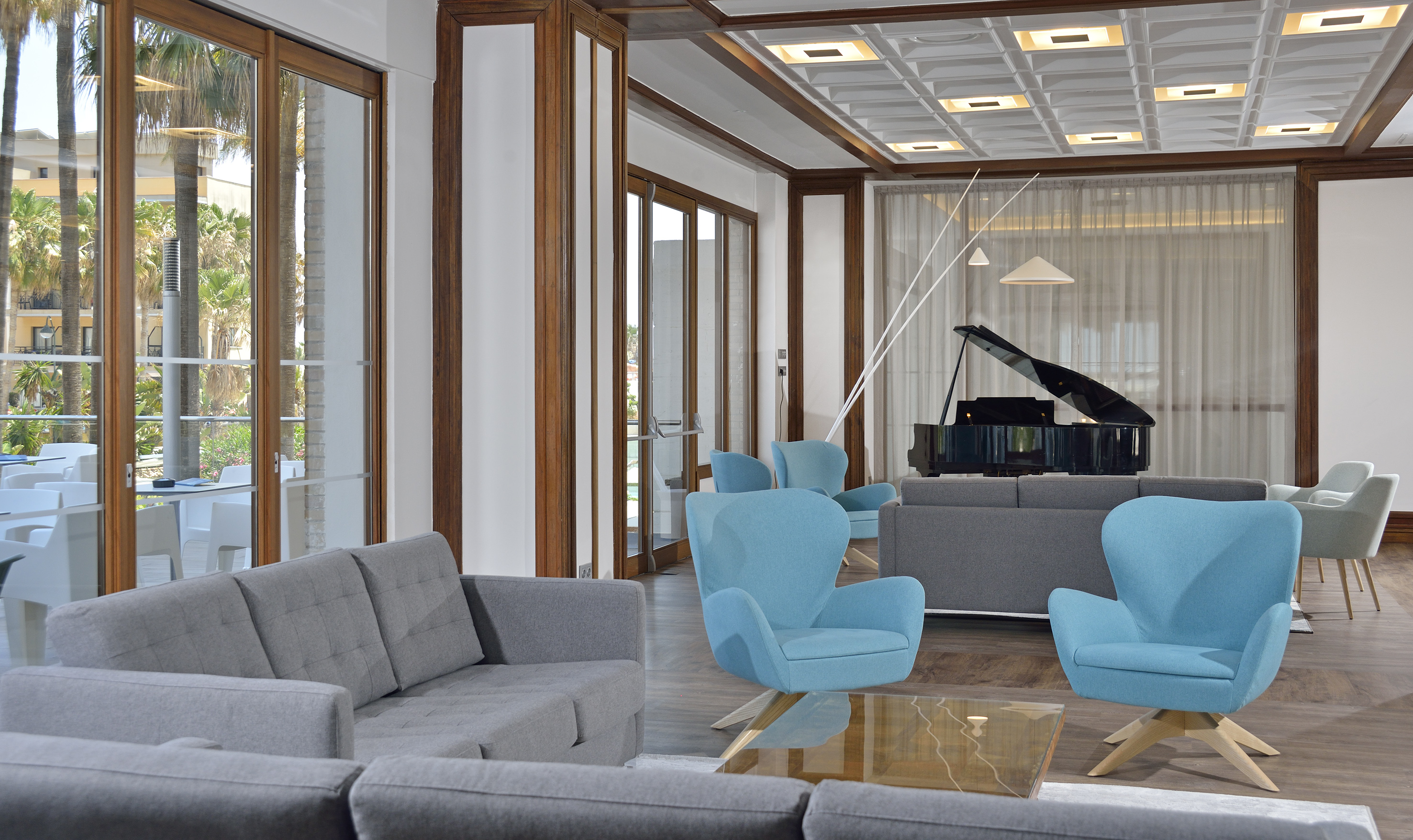 Modern hotel lounge: grey sofas, blue armchairs, grand piano, palm tree view.